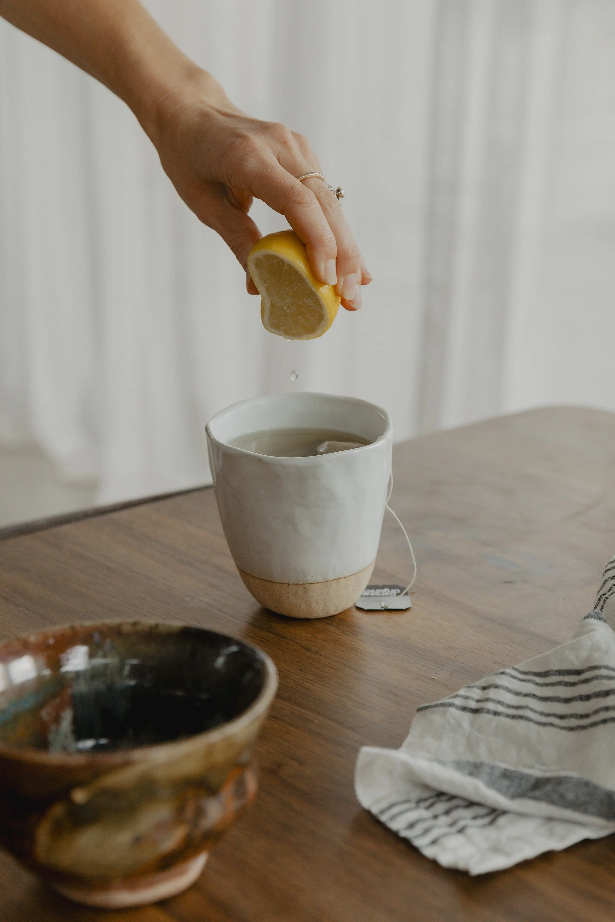 A hand squeezing a lemon into a white mug, with a tea bag on a wooden table and a striped cloth nearby.
