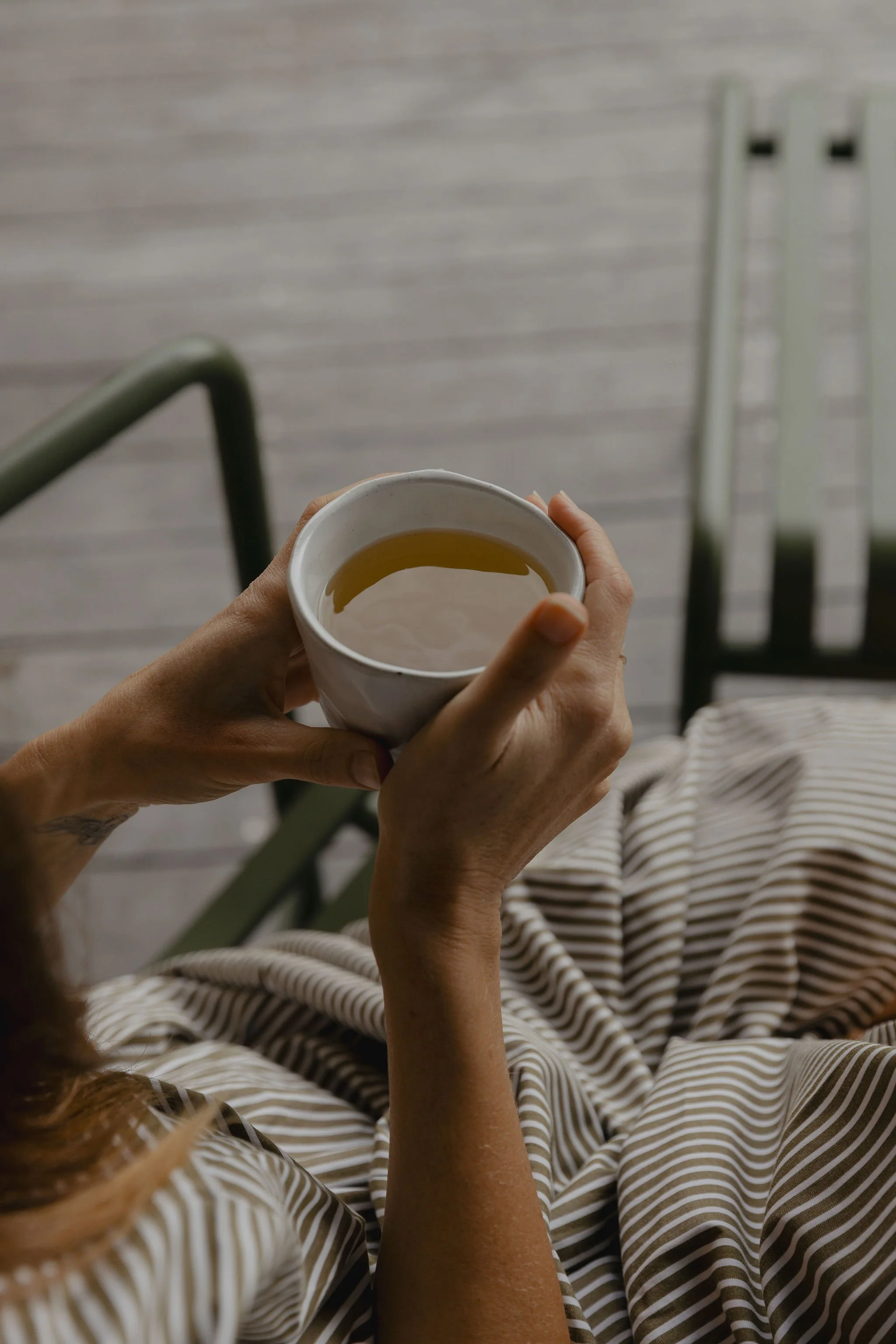 A person holding a white ceramic mug with a hot drink, sitting on a bed with striped bedding, near a gray wooden wall and railing.
