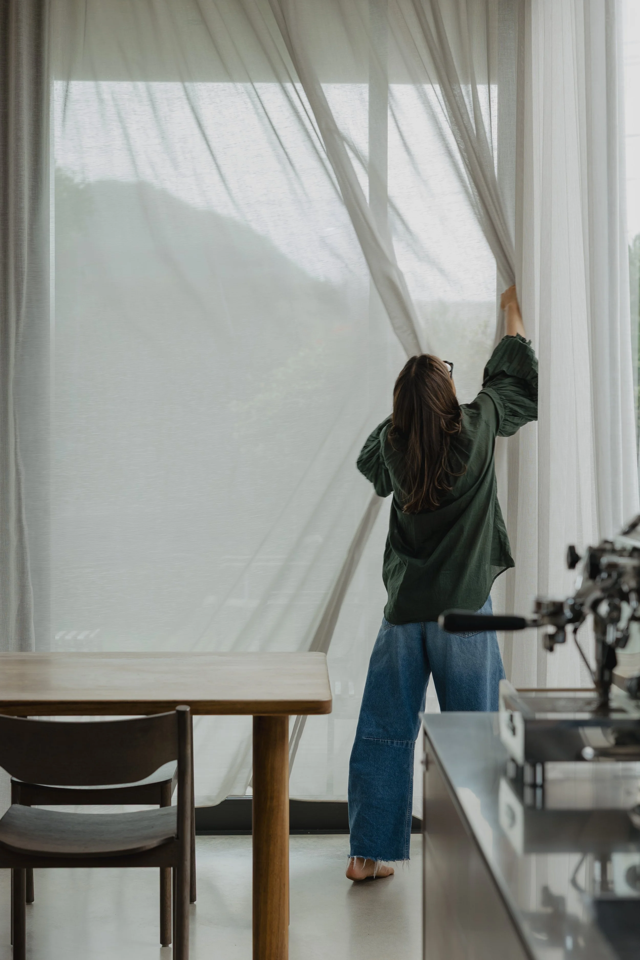A woman with brown hair, glasses, a green jacket, and jeans adjusting a white curtain in front of a large window in a room with a wooden dining table. It is daytime.
