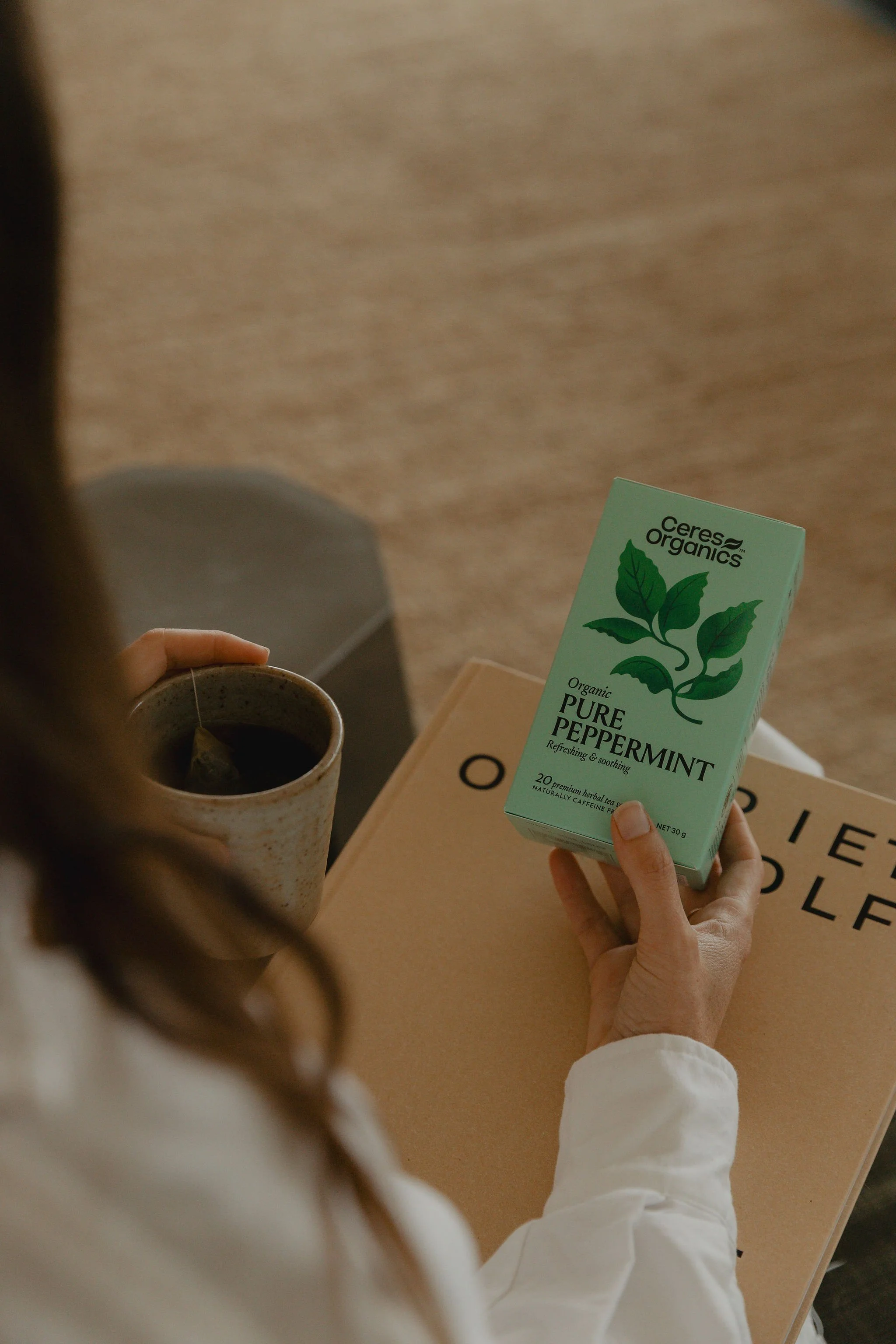 A person holding a box of Ceres Organics Pure Peppermint tea with a cup of tea nearby, sitting on a wooden table.