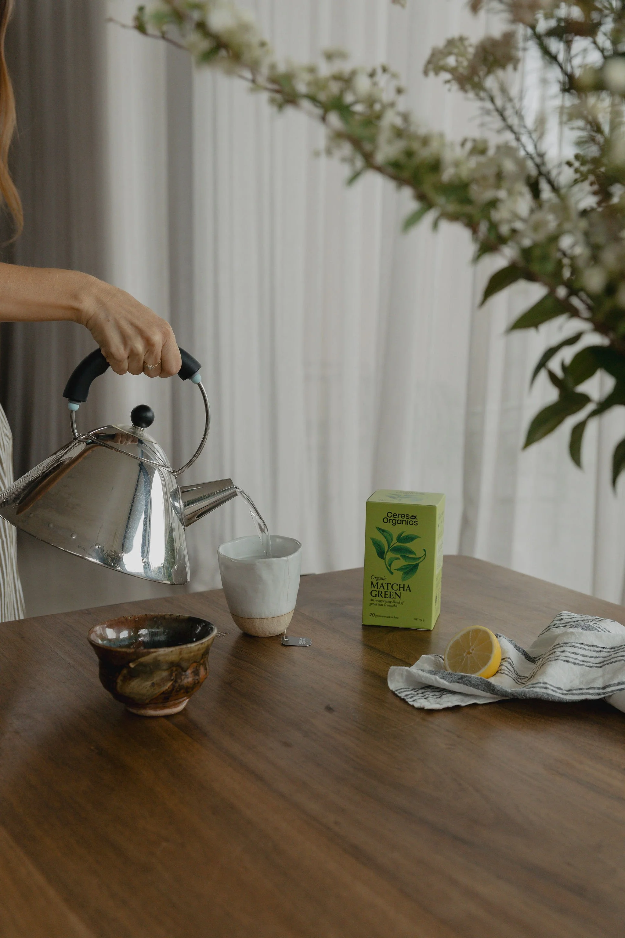 A person pouring hot water from a kettle into a white cup on a wooden table. On the table, there is a box of matcha green tea, a lemon, and two bowls. In the background, there are white curtains and a large flower arrangement.