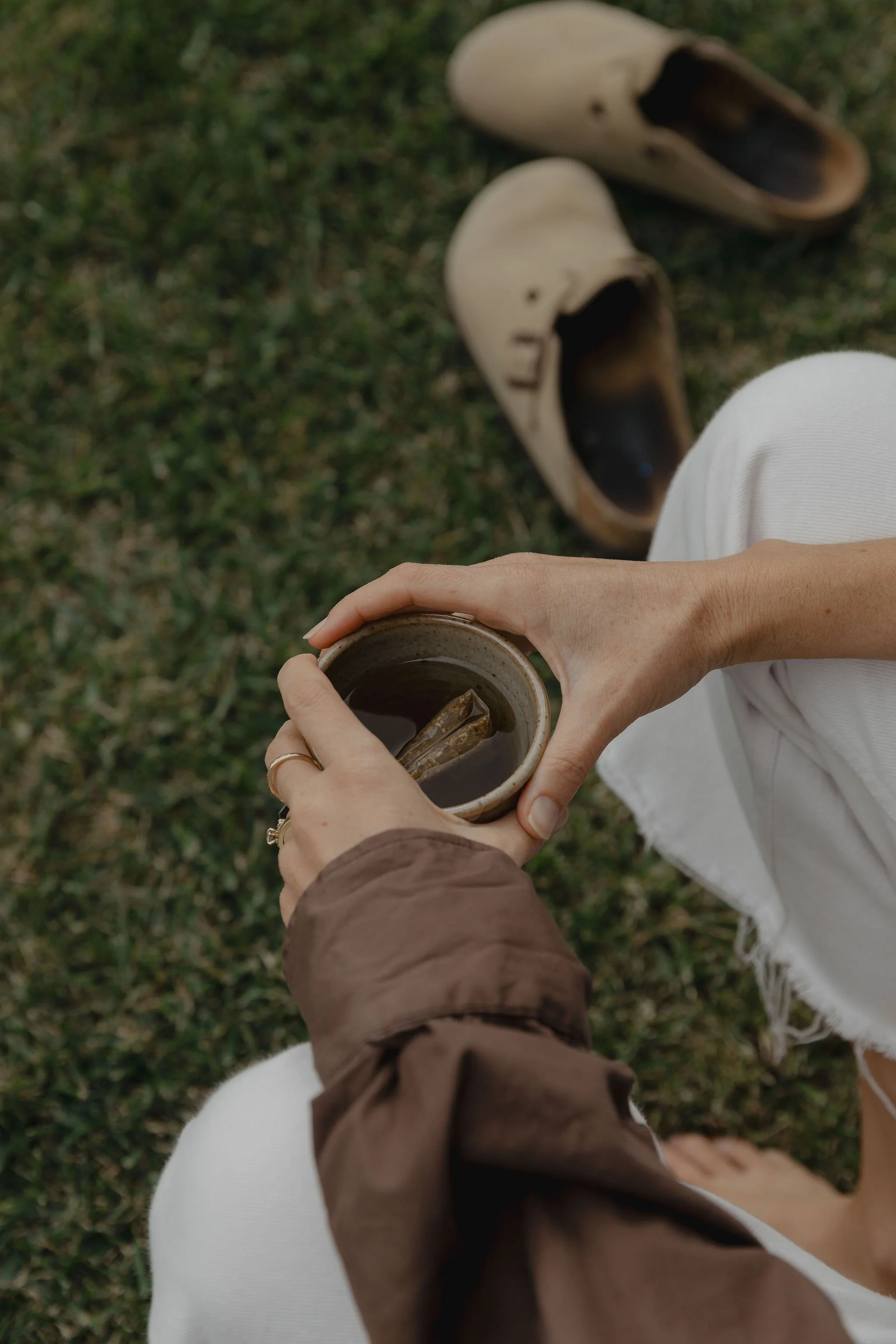 Overhead view of a person enjoying a cup of tea outdoors, with a pair of beige slip-on shoes on green grass in the background.