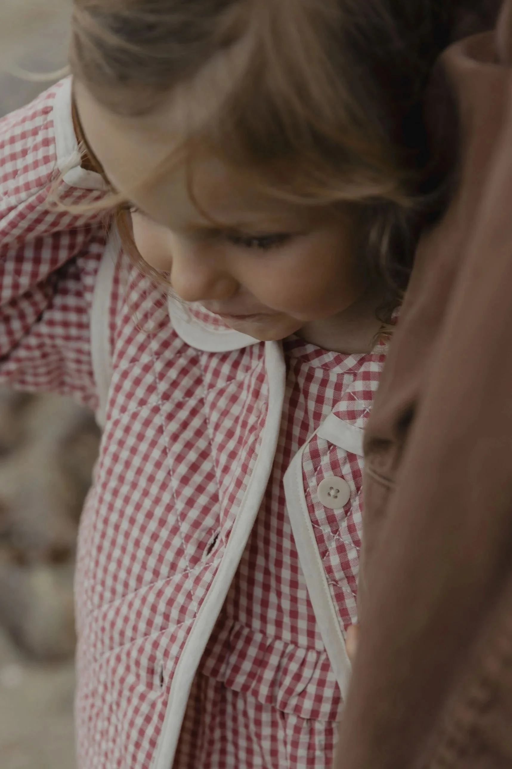 Close-up of a young child with light brown hair wearing a red and white checkered shirt, looking downward.