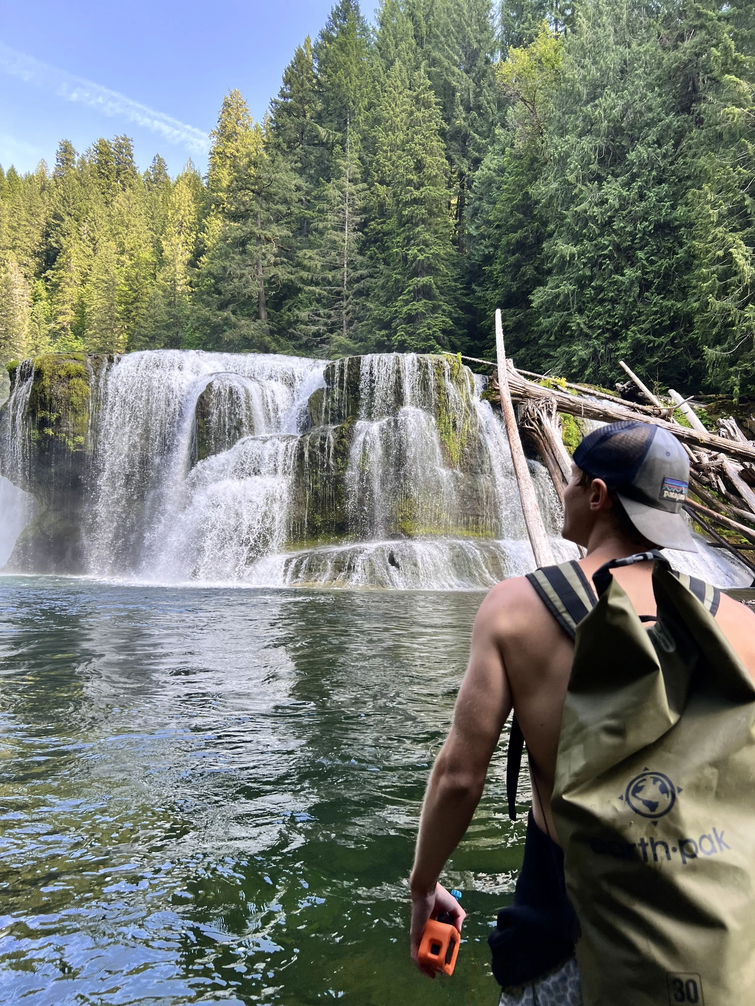A shirtless man with a backpack and cap looking at a waterfall in a forested area.