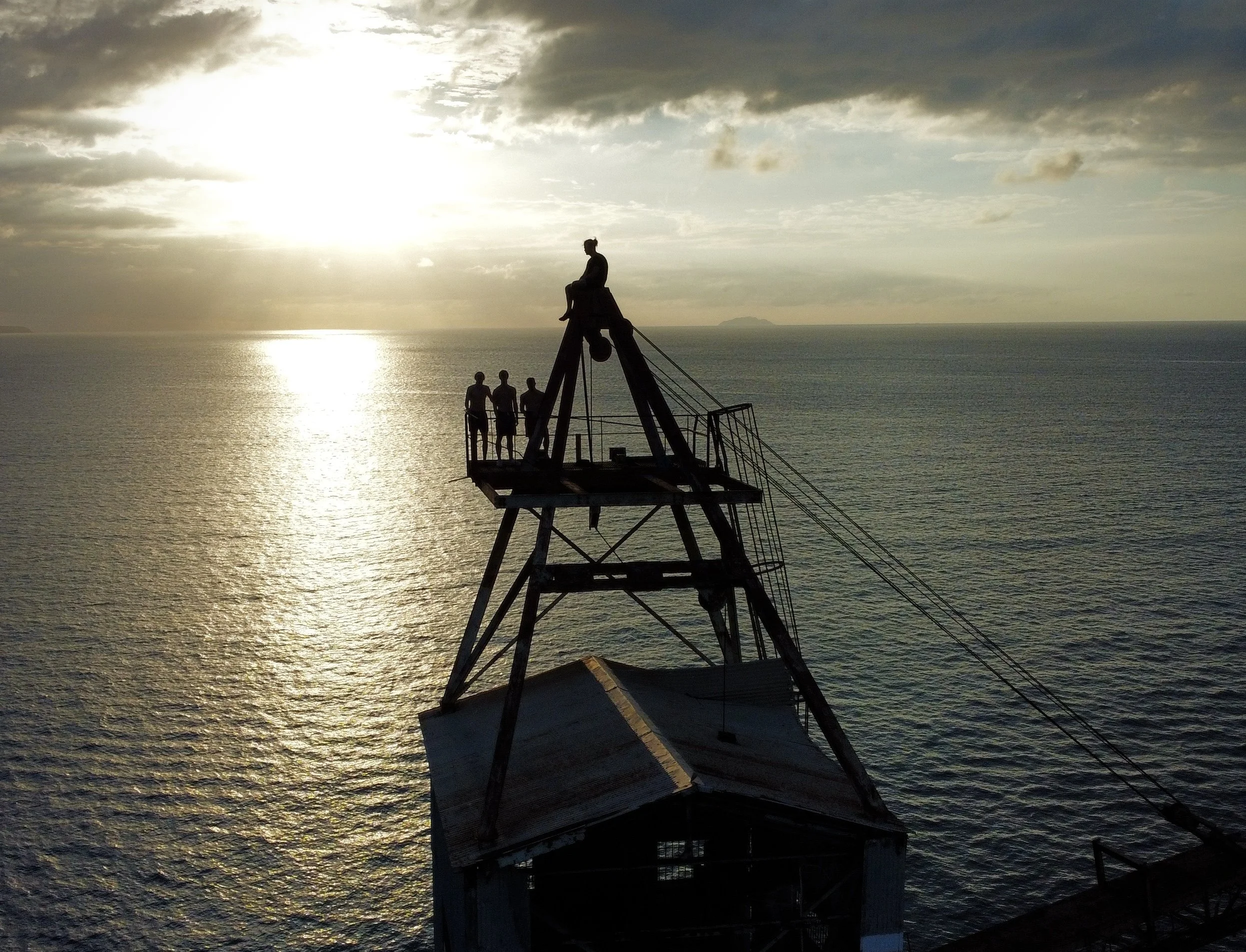 Silhouette of three people and one person sitting on a high point of a rusty metal crane structure overlooking the ocean at sunset or sunrise.
