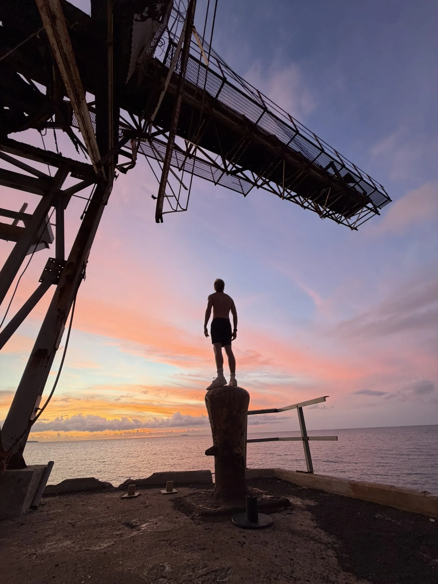 A person stands on a large military-style anchor on a coastline during a colorful sunset, with a tall metal structure (possibly an old crane or tower) nearby.
