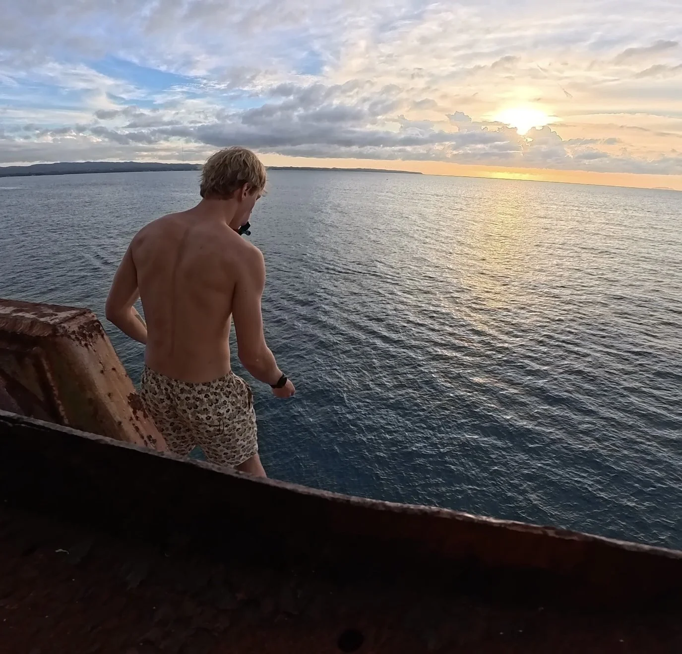 A shirtless young man with blonde hair wearing leopard print shorts standing on a rusty metal structure by the water during sunset.