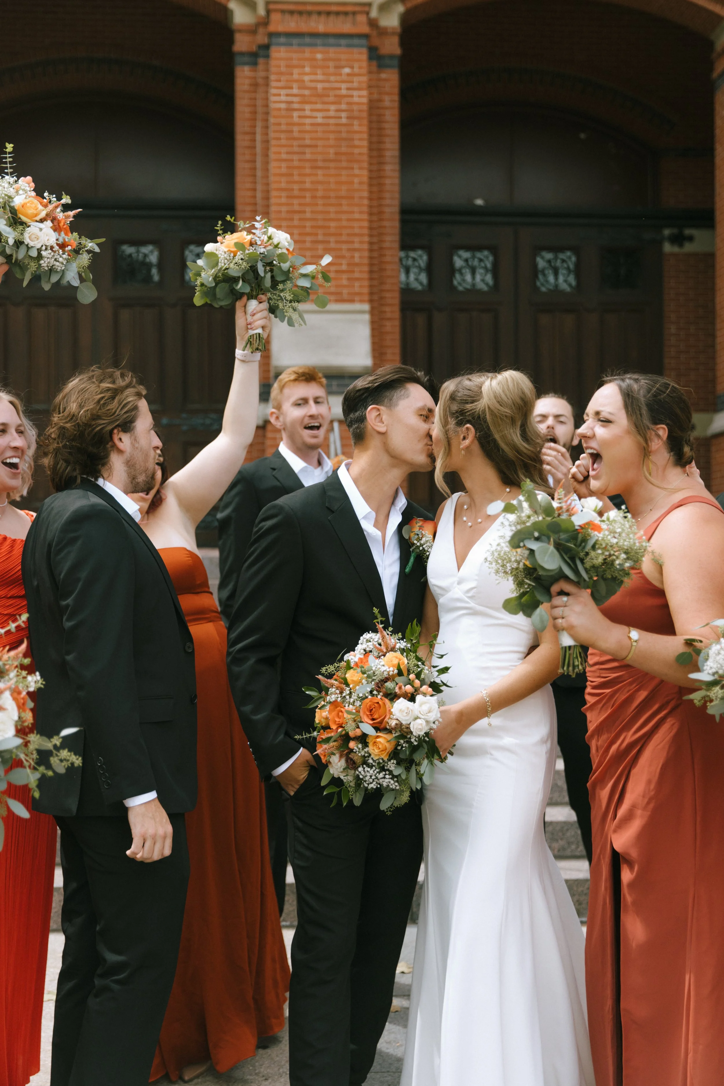 A wedding scene featuring a bride and groom kissing surrounded by wedding party members, with women in matching rust-colored dresses and men in black tuxedos, holding bouquets of flowers, in front of a brick building.