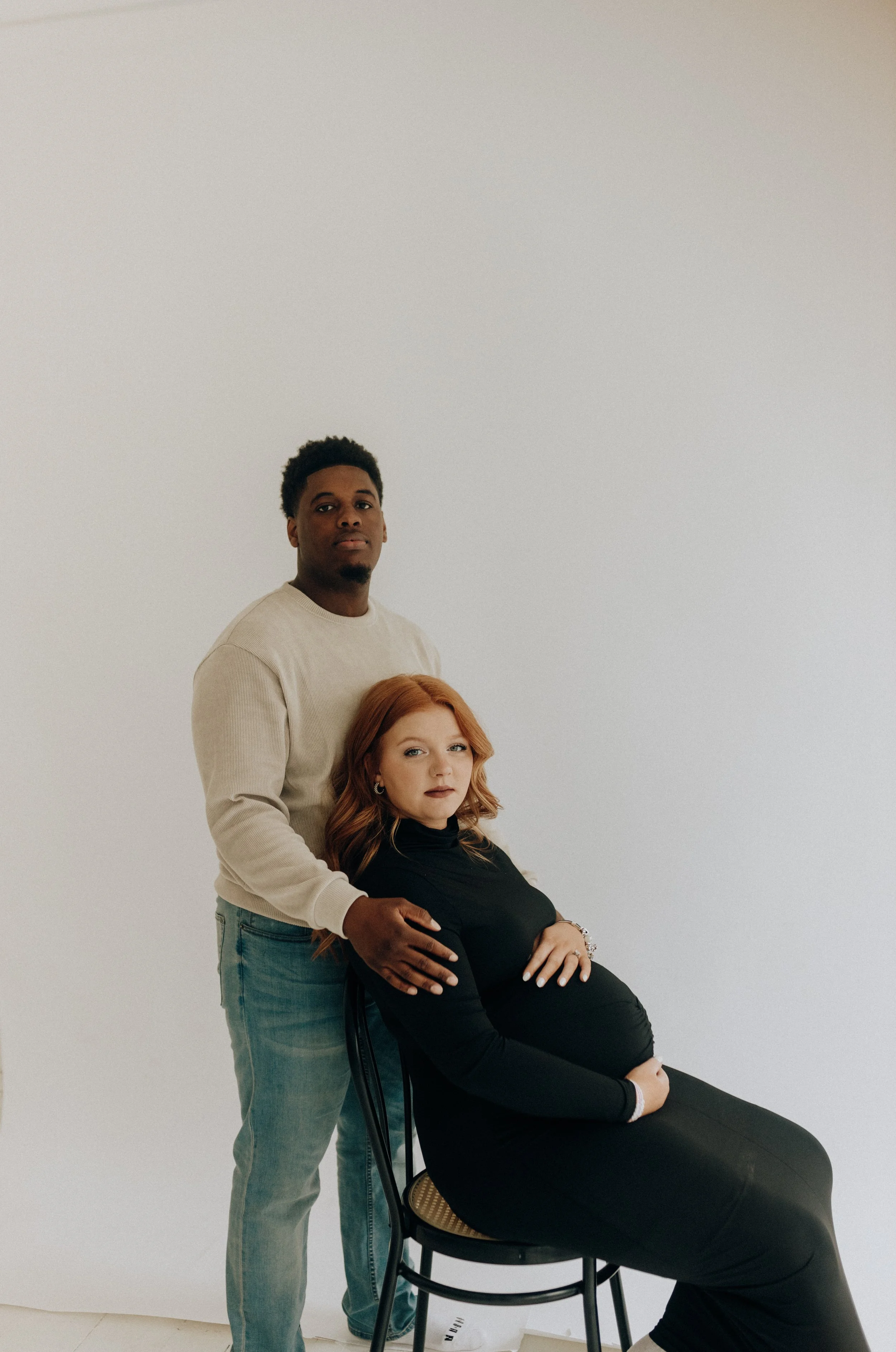 A pregnant woman with long red hair seated on a chair, with a man standing behind her, resting his hand on her shoulder. They are in a plain, light-colored room.
