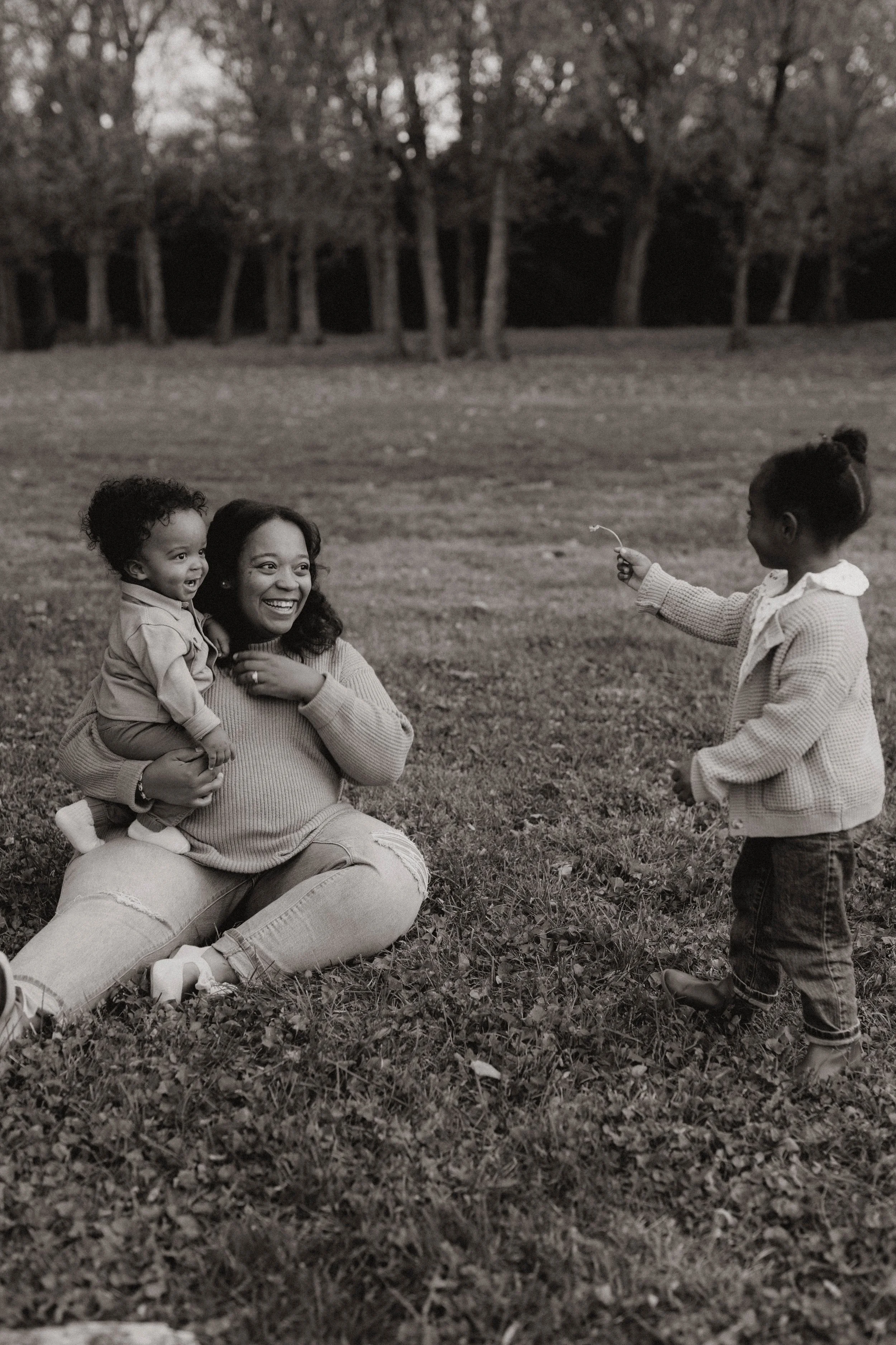 A woman sitting on the grass holding a young child, while another child is standing nearby holding a dandelion outdoors in a park.