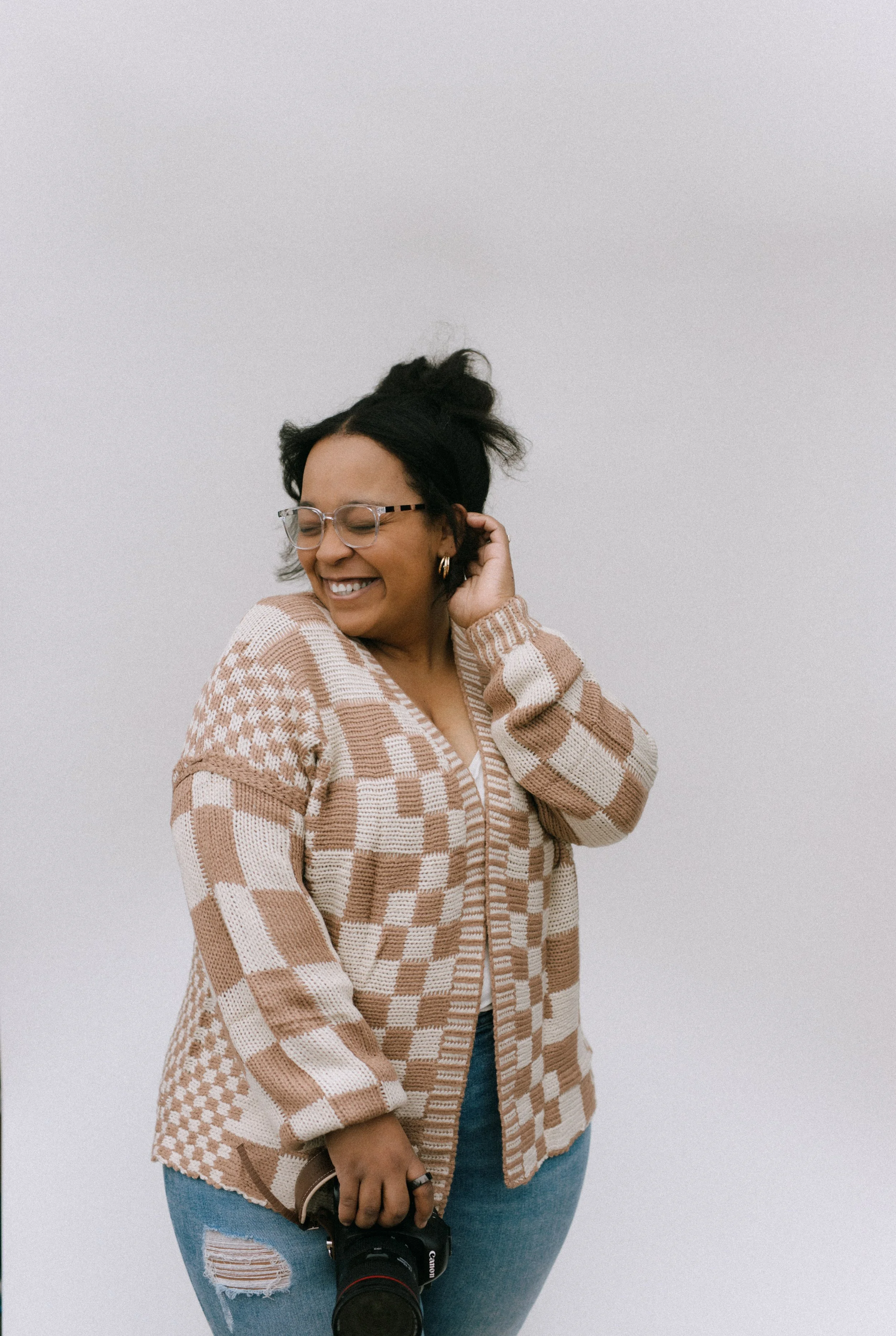 A woman with glasses and earrings, smiling with eyes closed, wearing a patterned beige and white cardigan, holding a camera, standing against a plain light background.