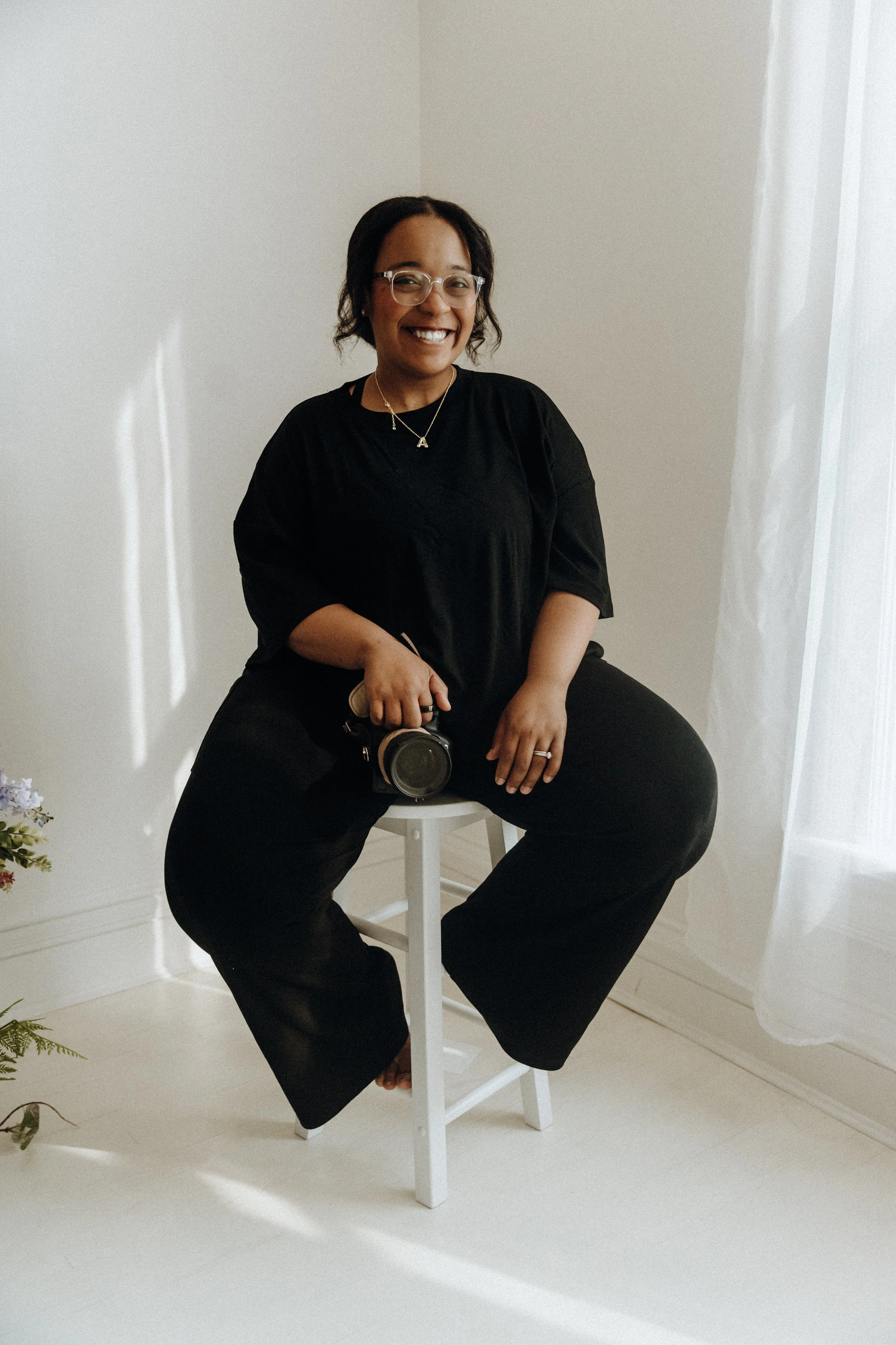 A woman wearing glasses, a black shirt, and black pants, sitting on a white stool, holding a camera, smiling in a well-lit room with white walls and curtains.