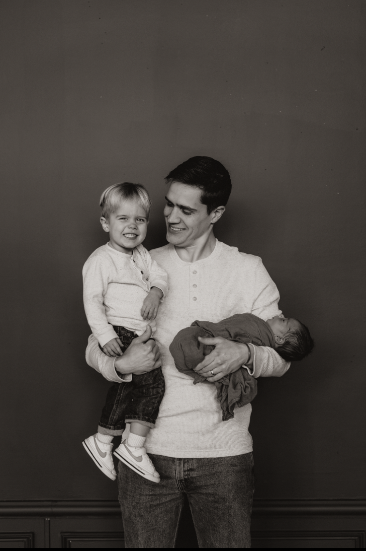A man holding a young boy in his left arm and a newborn baby in his right arm, standing against a plain dark background.