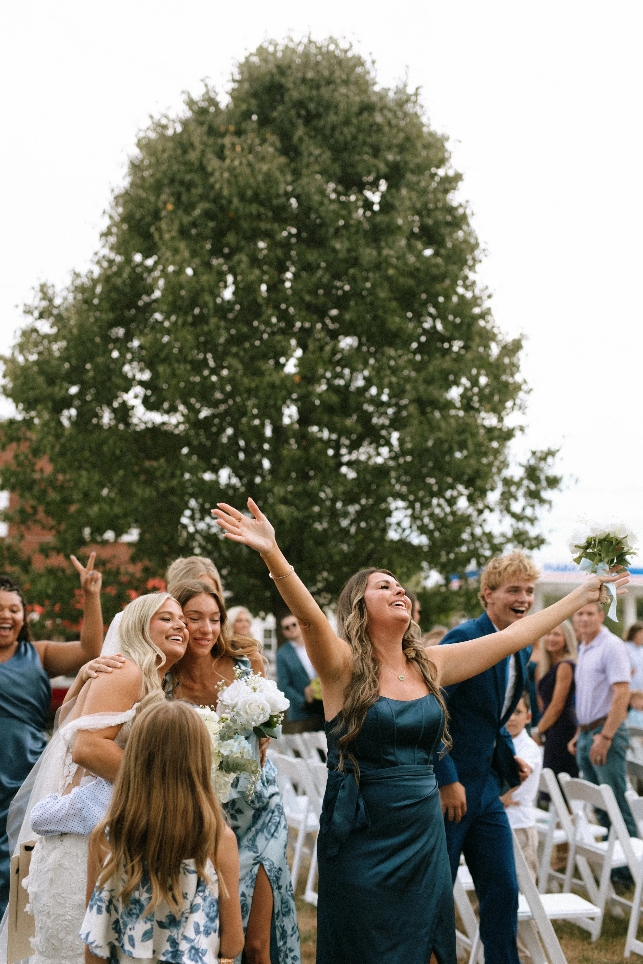Group of people celebrating at an outdoor wedding, with a woman in a dark dress holding a bouquet and raising her arms in joy, and others smiling and dancing around her, set against a large tree and chairs.
