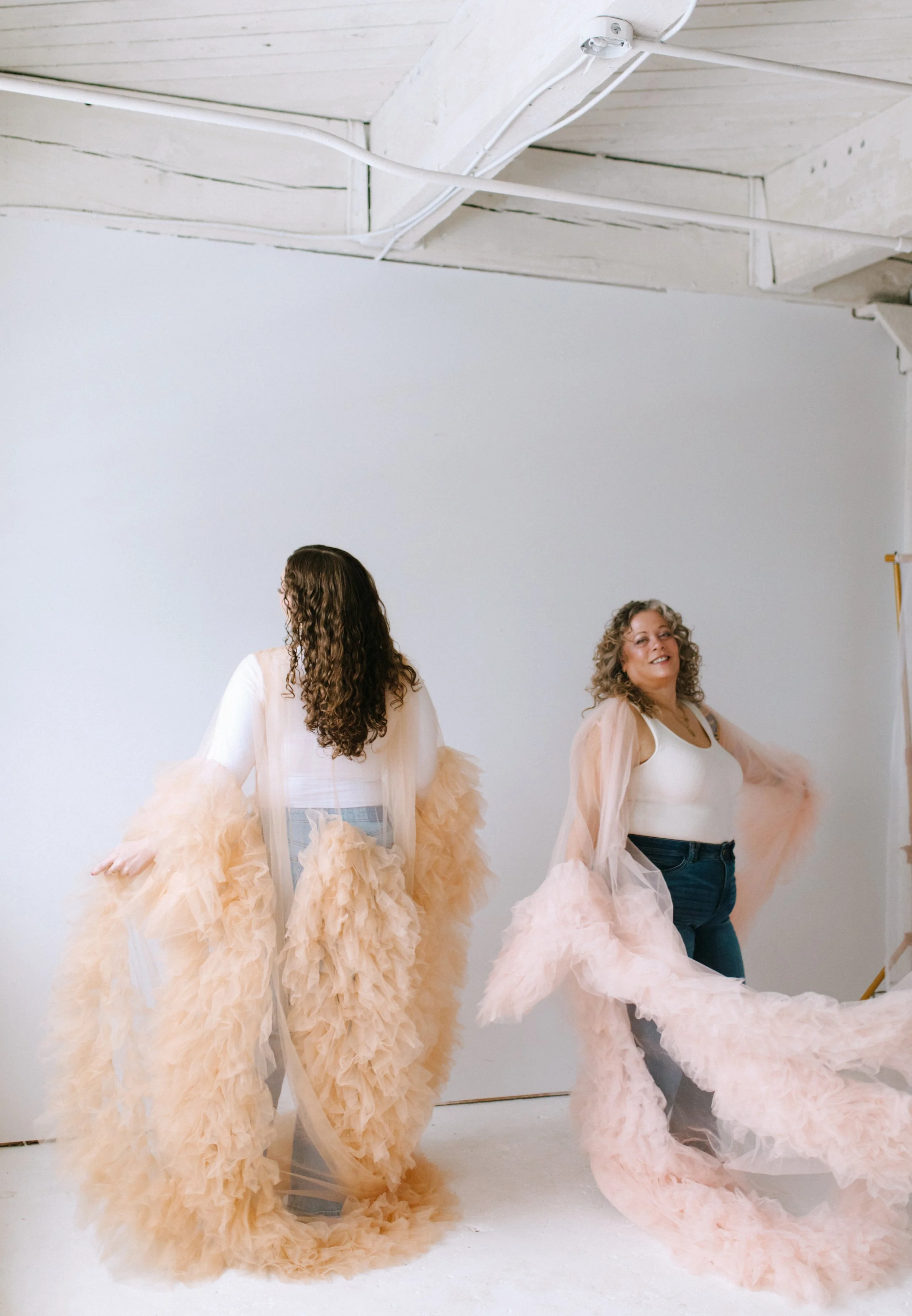 Two women posing in a studio, wearing luxurious, peach-colored, ruffled, sheer robes over casual clothing. The woman on the right is smiling and has curly hair, while the woman on the left is facing away.