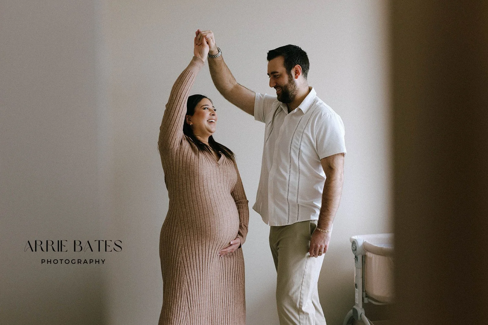 A joyful pregnant woman in a brown dress dancing with a smiling man in a white shirt, in a softly lit room.