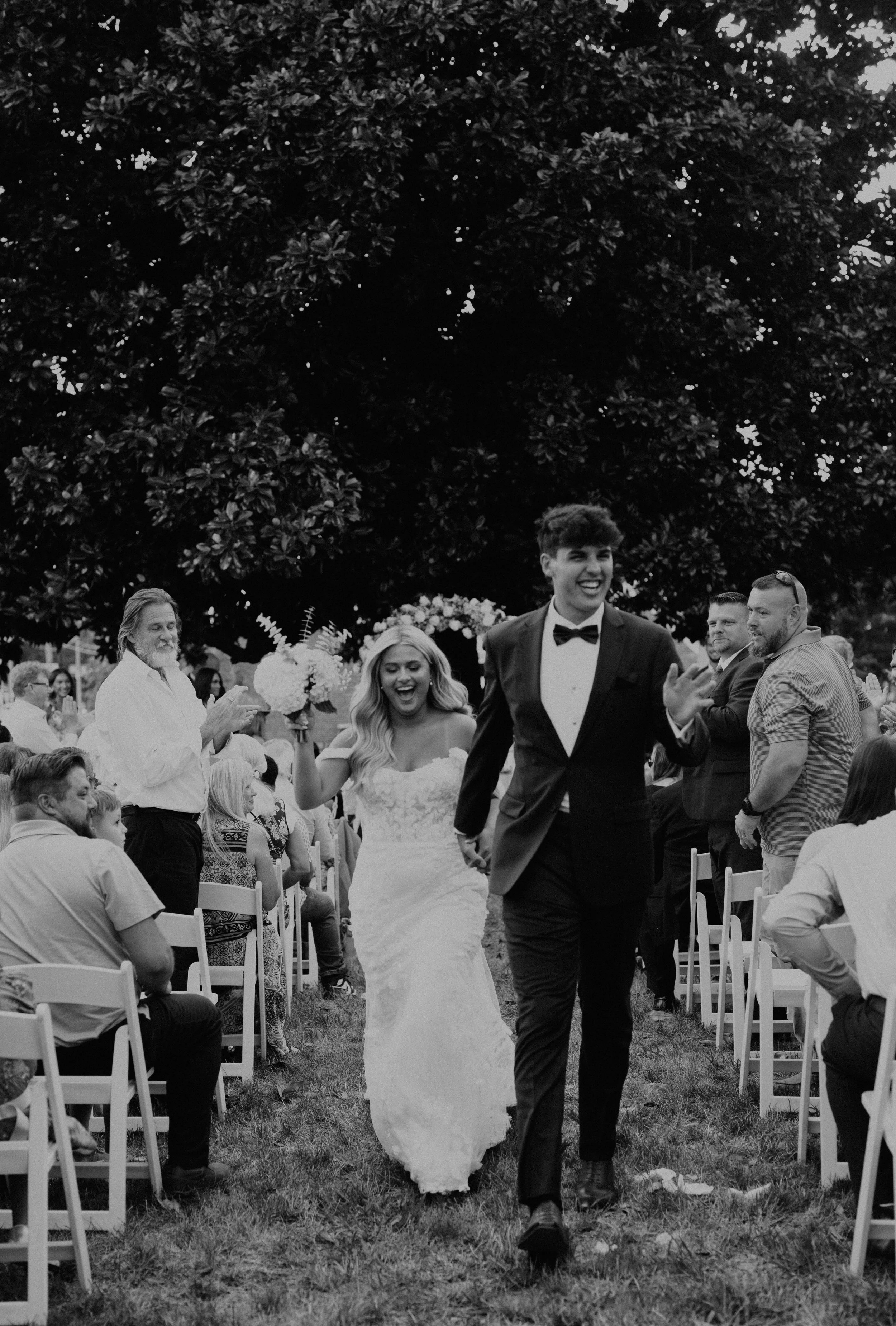 A black and white photo of a wedding ceremony outdoors with a bride and groom walking down the aisle, surrounded by guests seated on both sides, and a large tree overhead.