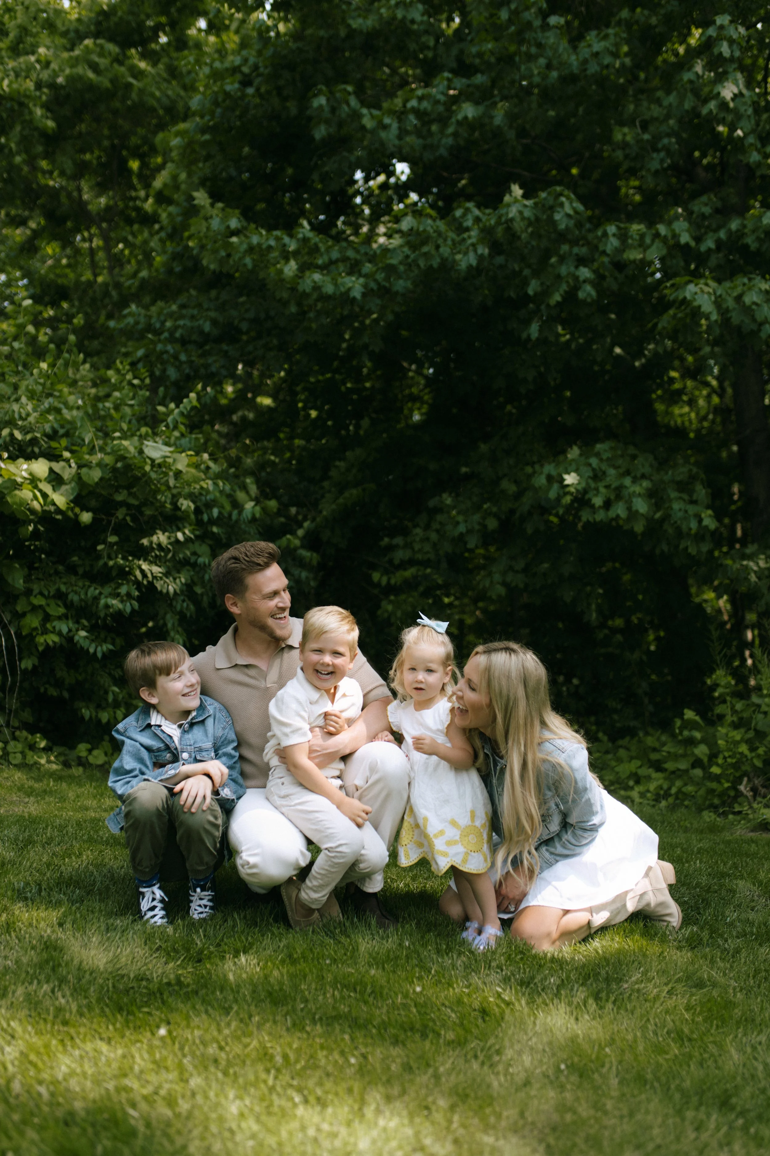 A family of five, including two adult women and three children, joyfully posing on a grassy lawn with green trees in the background.