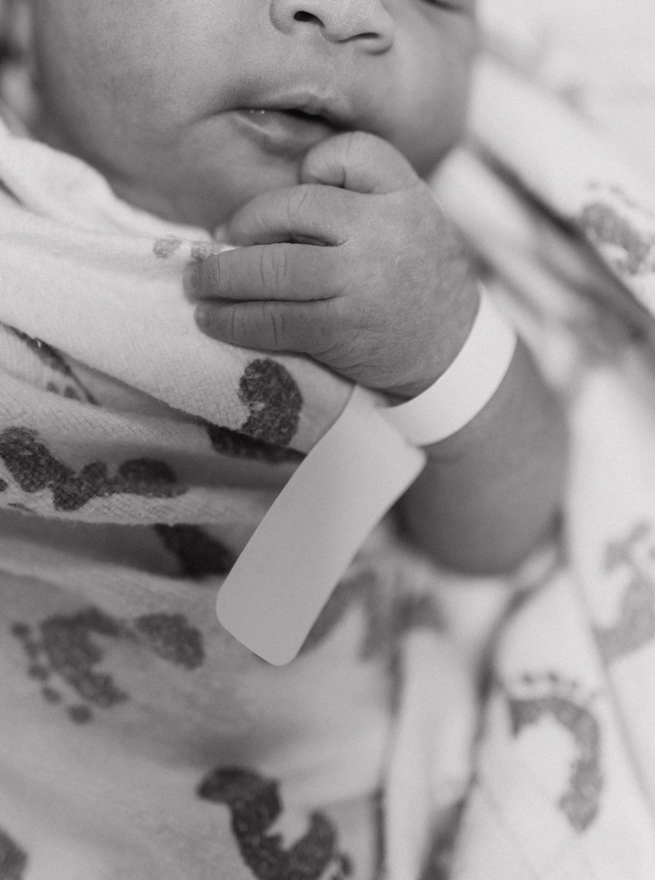 Close-up of a newborn baby's hand holding a finger, with hospital identification bracelet and patterned clothing visible, in black and white.