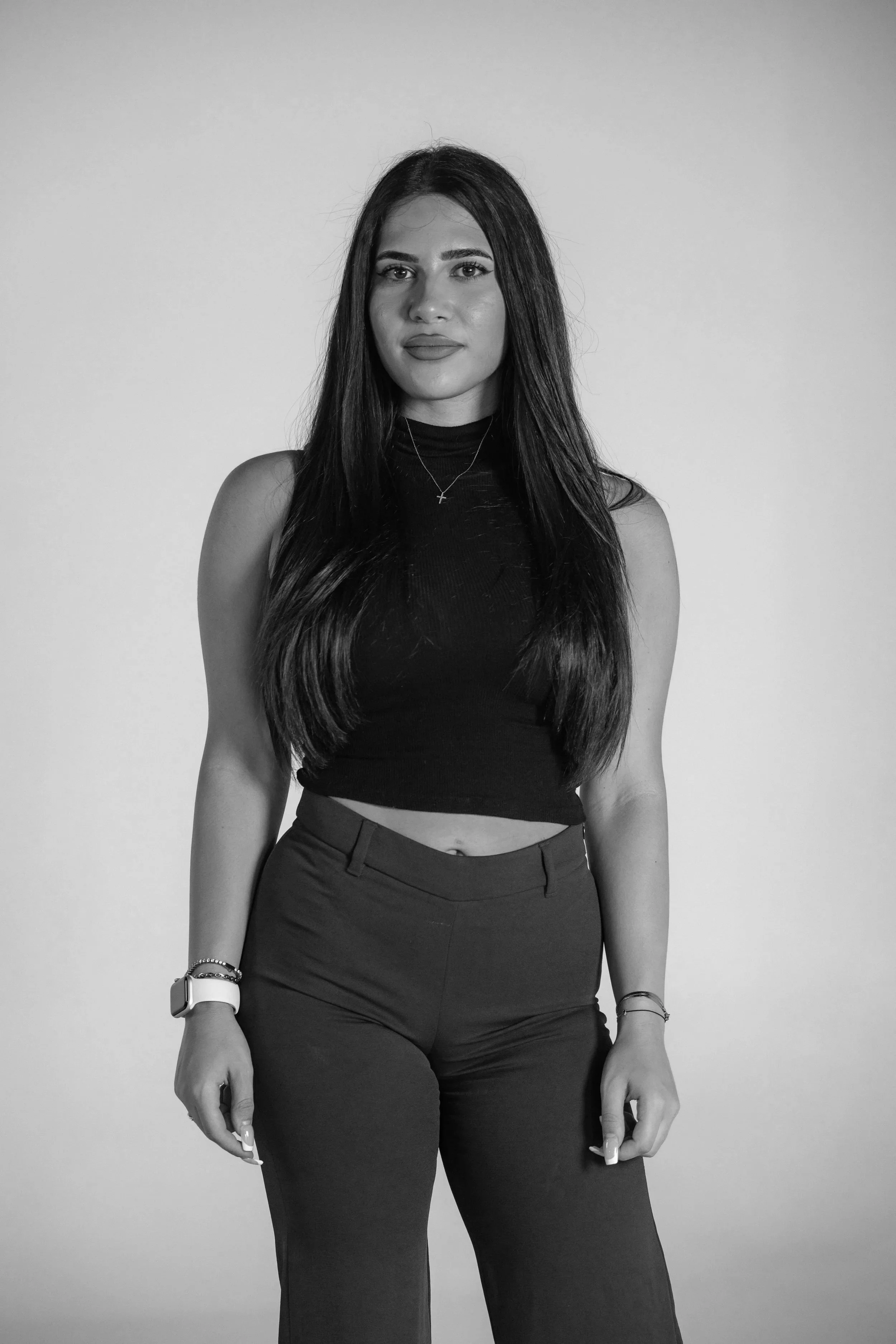 Black and white photo of a young woman with long dark hair, wearing a sleeveless top and high-waisted pants, standing against a plain background.