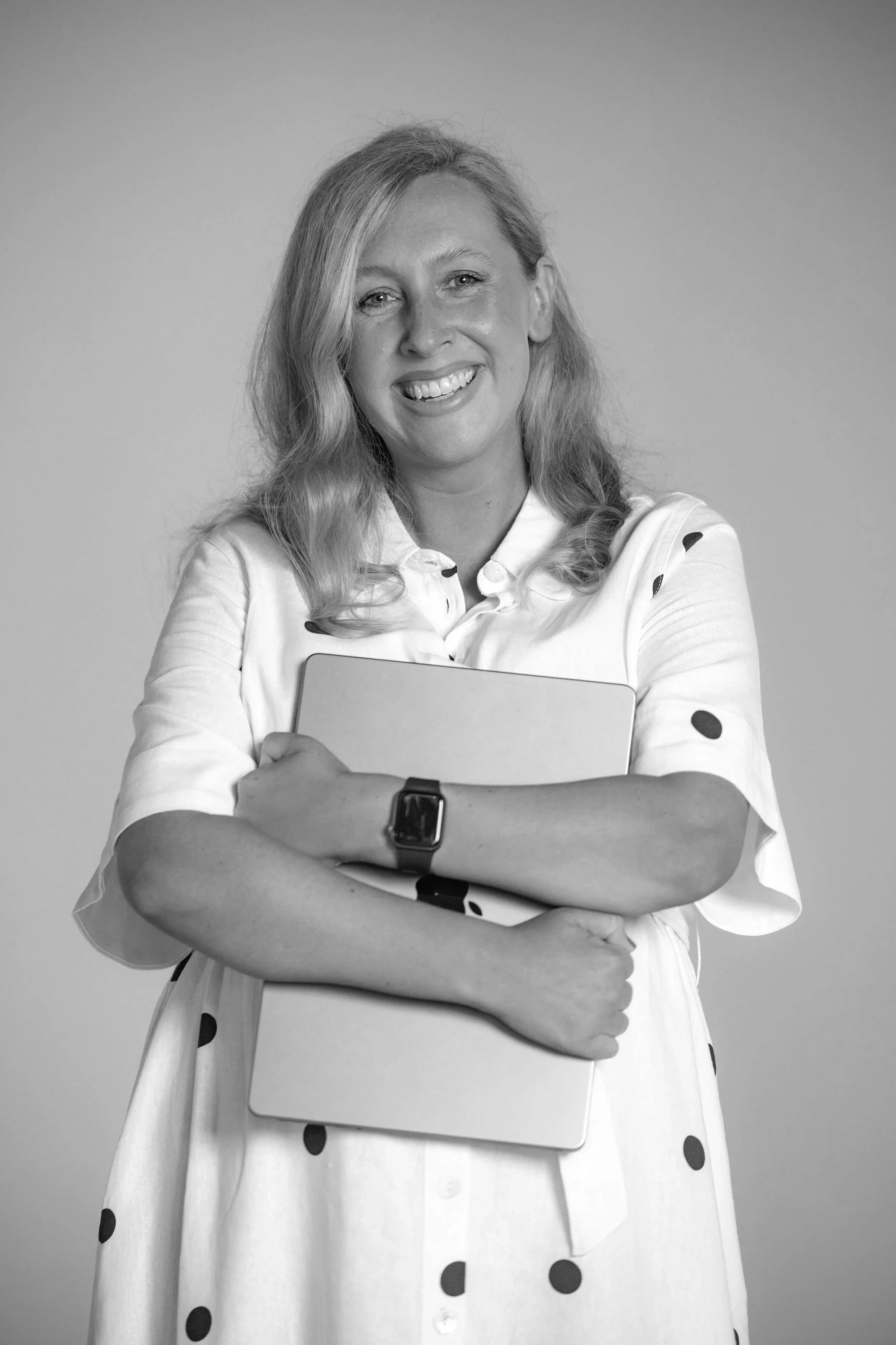 Black and white photo of a smiling woman with wavy hair, wearing a polka dot dress, holding a closed laptop, with her arms crossed, wearing a smartwatch.