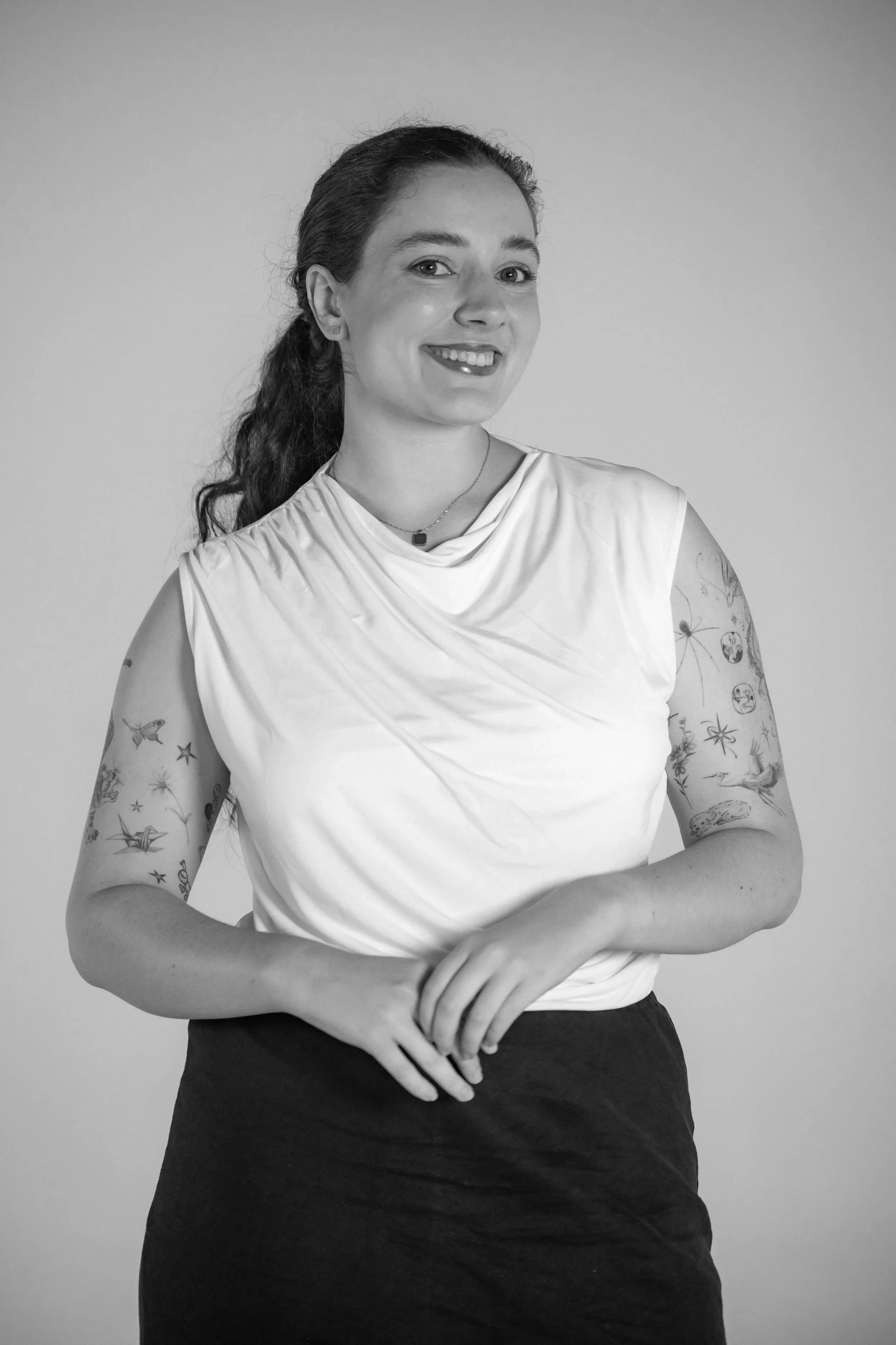 Black and white portrait of a young woman with tattoos on both arms, smiling and looking at the camera, wearing a sleeveless white top and a necklace.