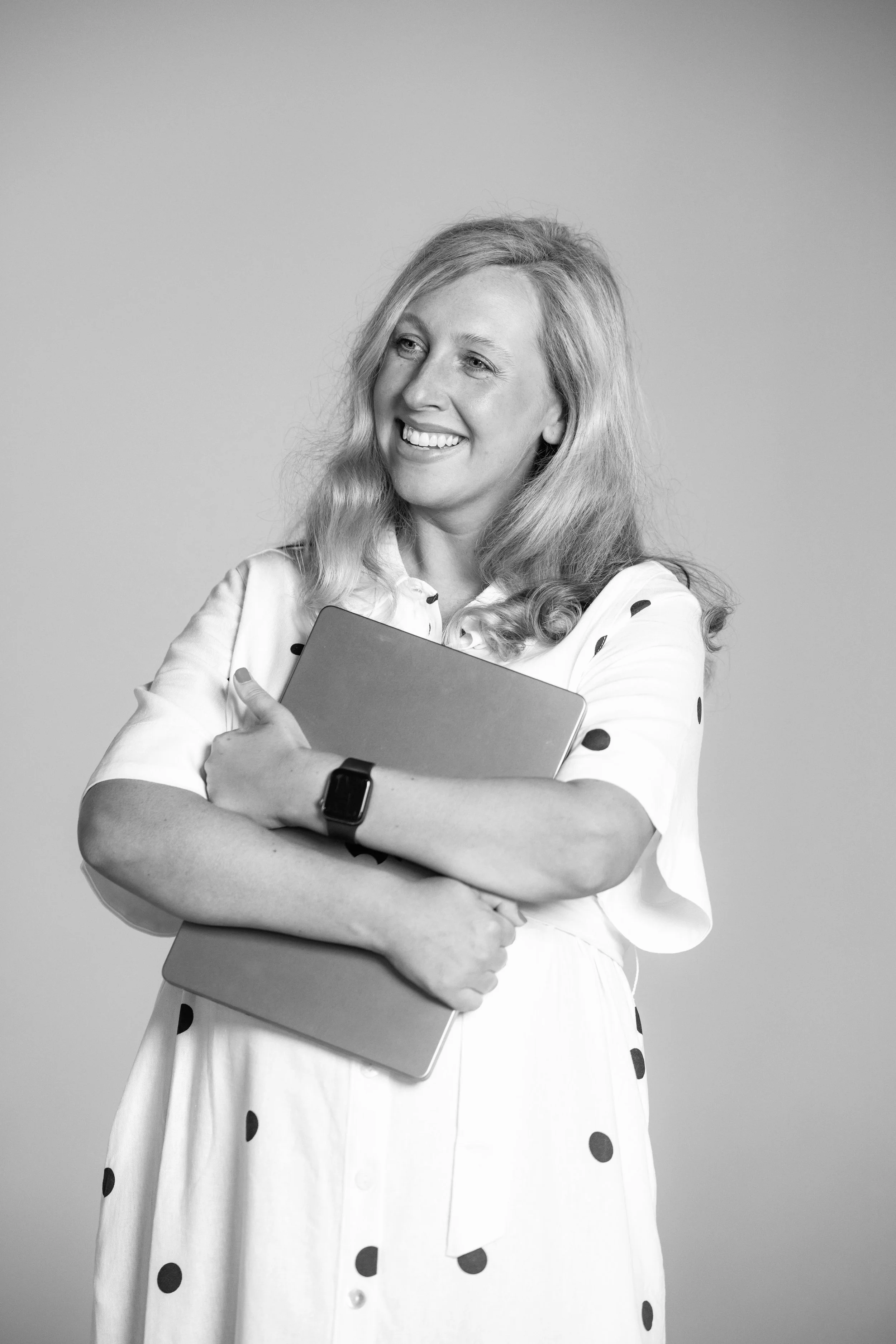 Black and white photo of Emily Cooke, Founder & Director of Brandshake, smiling and looking to her left, holding a tablet or notebook in front of her. She is wearing a white dress with black polka dots and a smartwatch on her left wrist.
