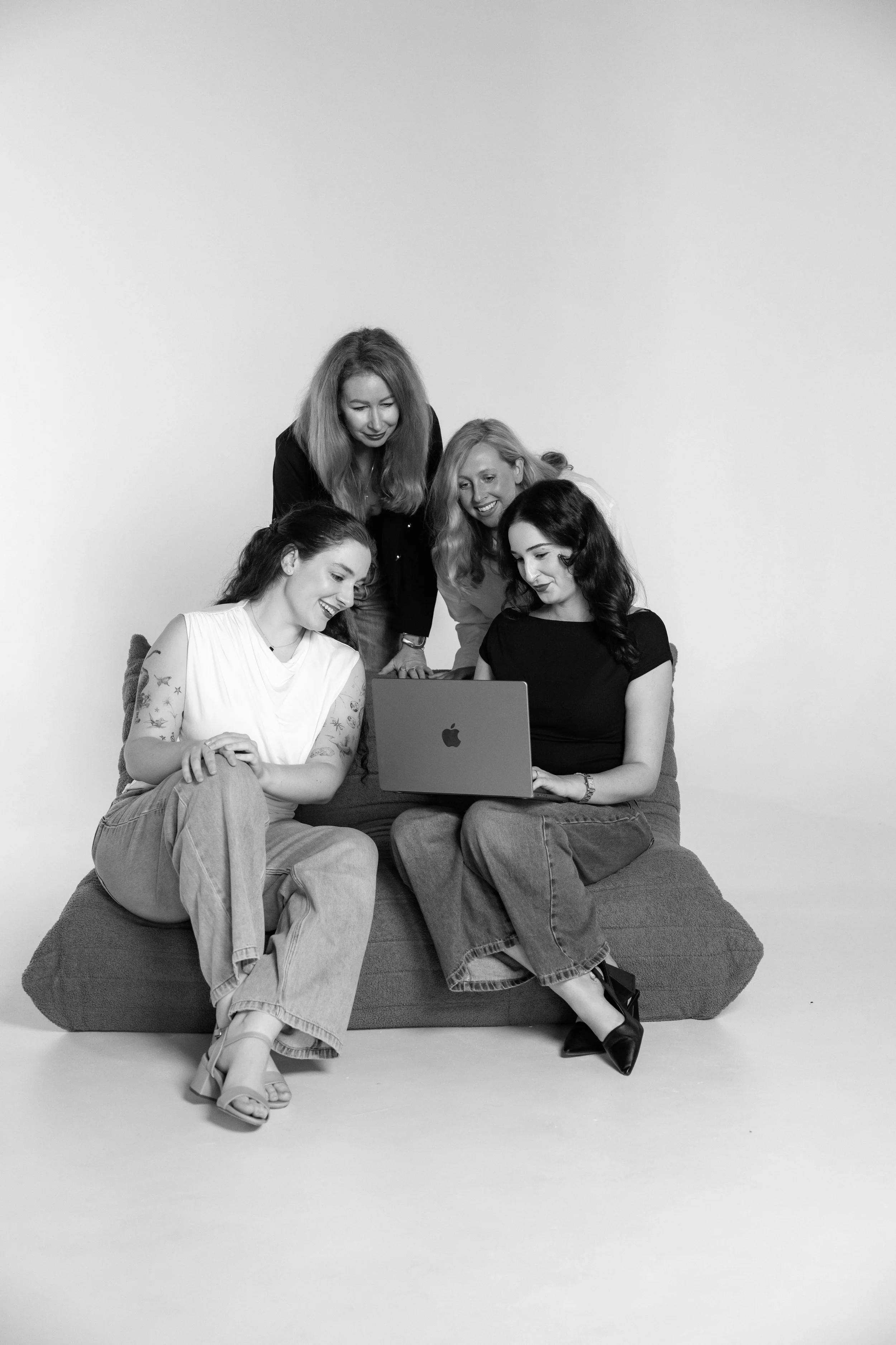 Team Brandshake (Em, Gabby, Lyn and Christina) gathered around a laptop, sitting on a bean bag chair, with one woman standing behind them, all smiling and looking at the laptop. All are casually dressed and appear to be having a discussion.
