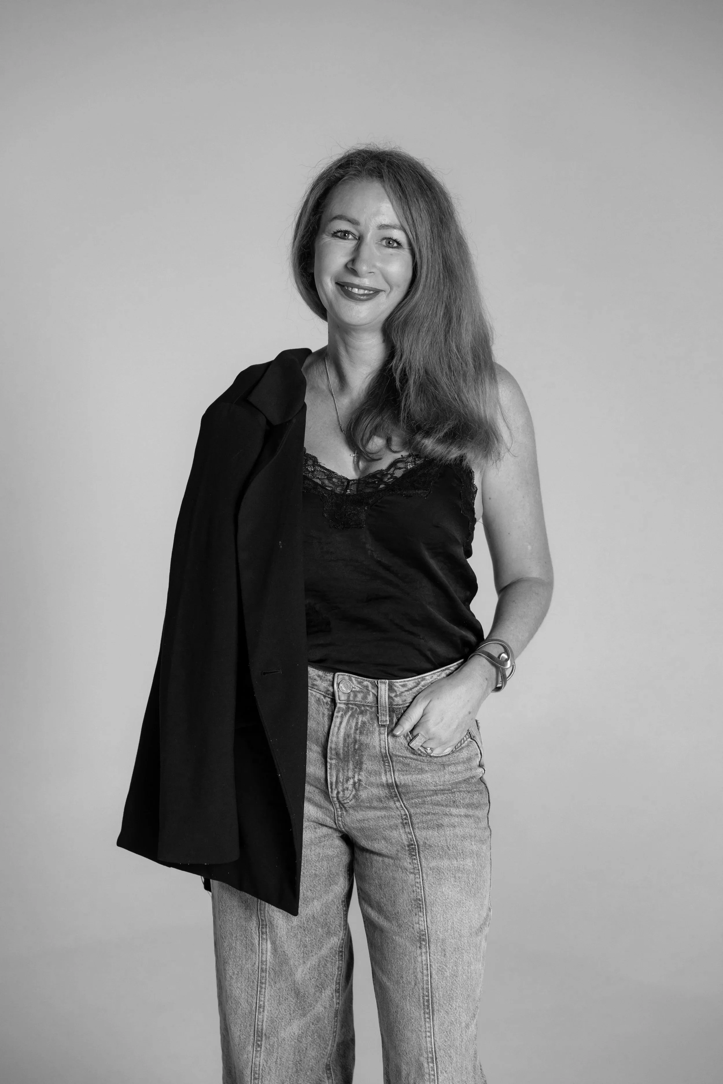 A woman with long hair, wearing a black lace top and jeans, holding a jacket over her shoulder, smiling for the camera.