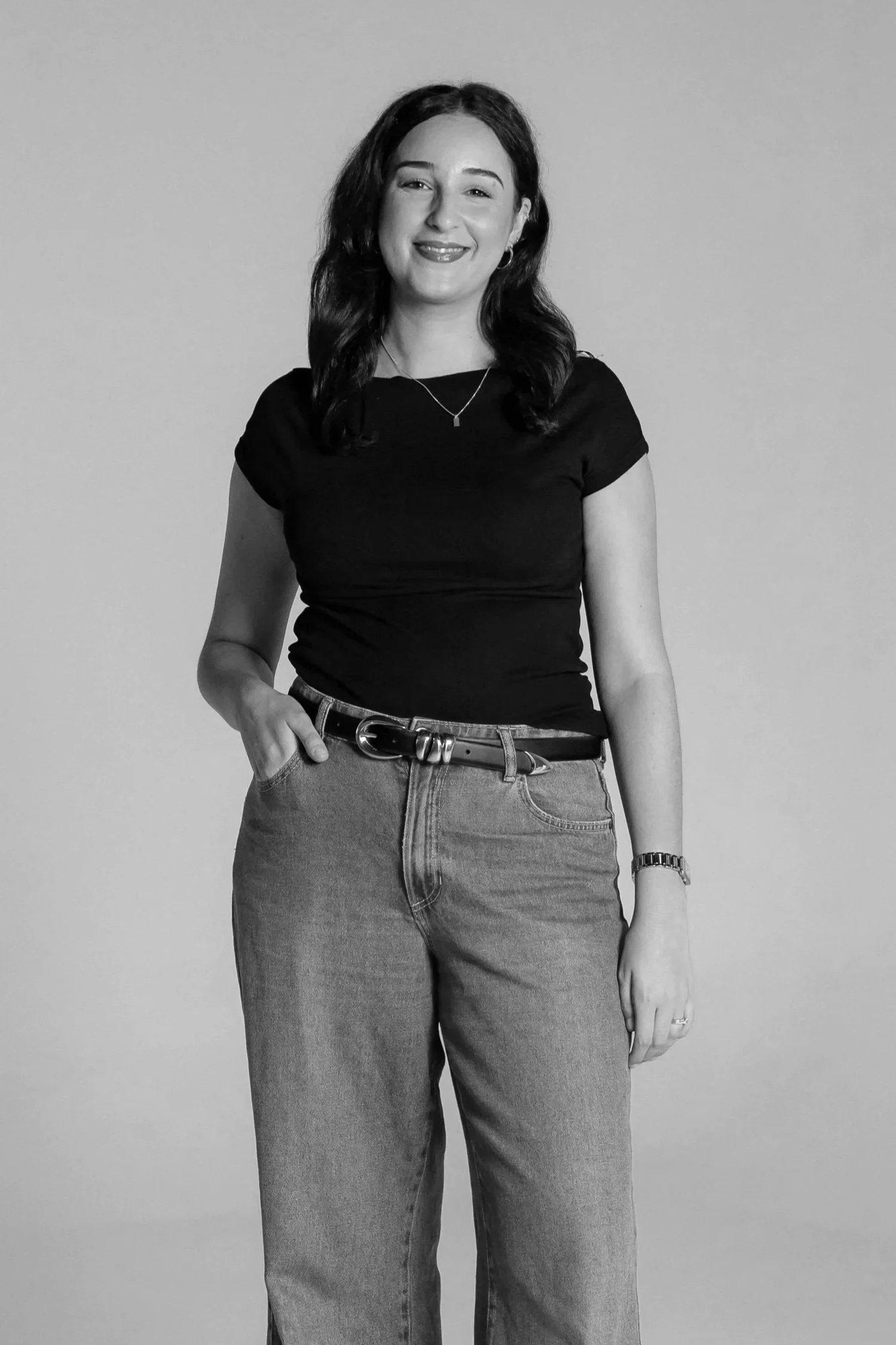 Black and white photo of a woman with dark, wavy hair wearing a black t-shirt, high-waisted jeans, and a watch, standing against a plain background, smiling with one hand in her pocket.
