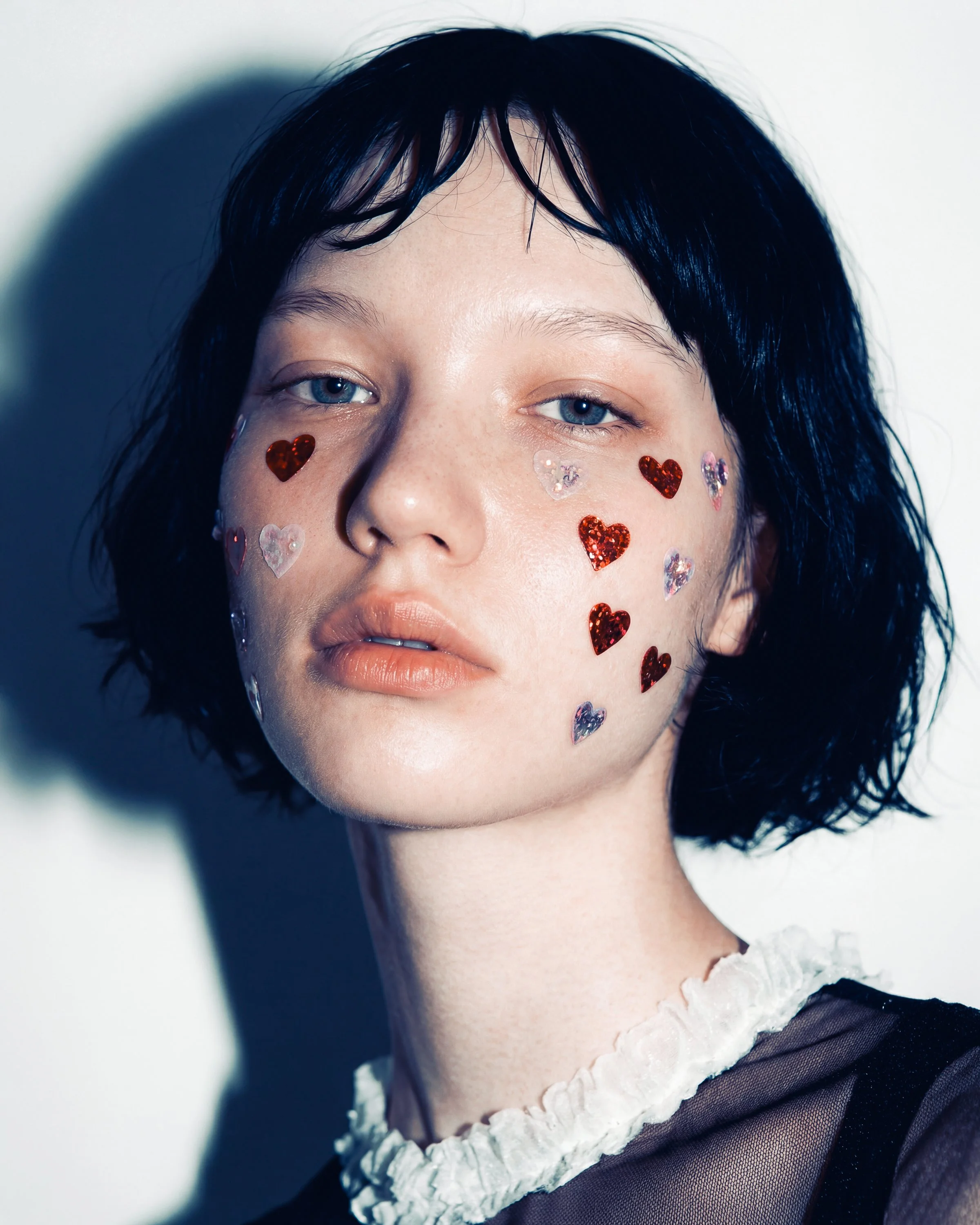 Portrait of a woman with short black hair and blue eyes, decorated with red and translucent heart-shaped stickers on her face, wearing a black top with a ruffled white collar.