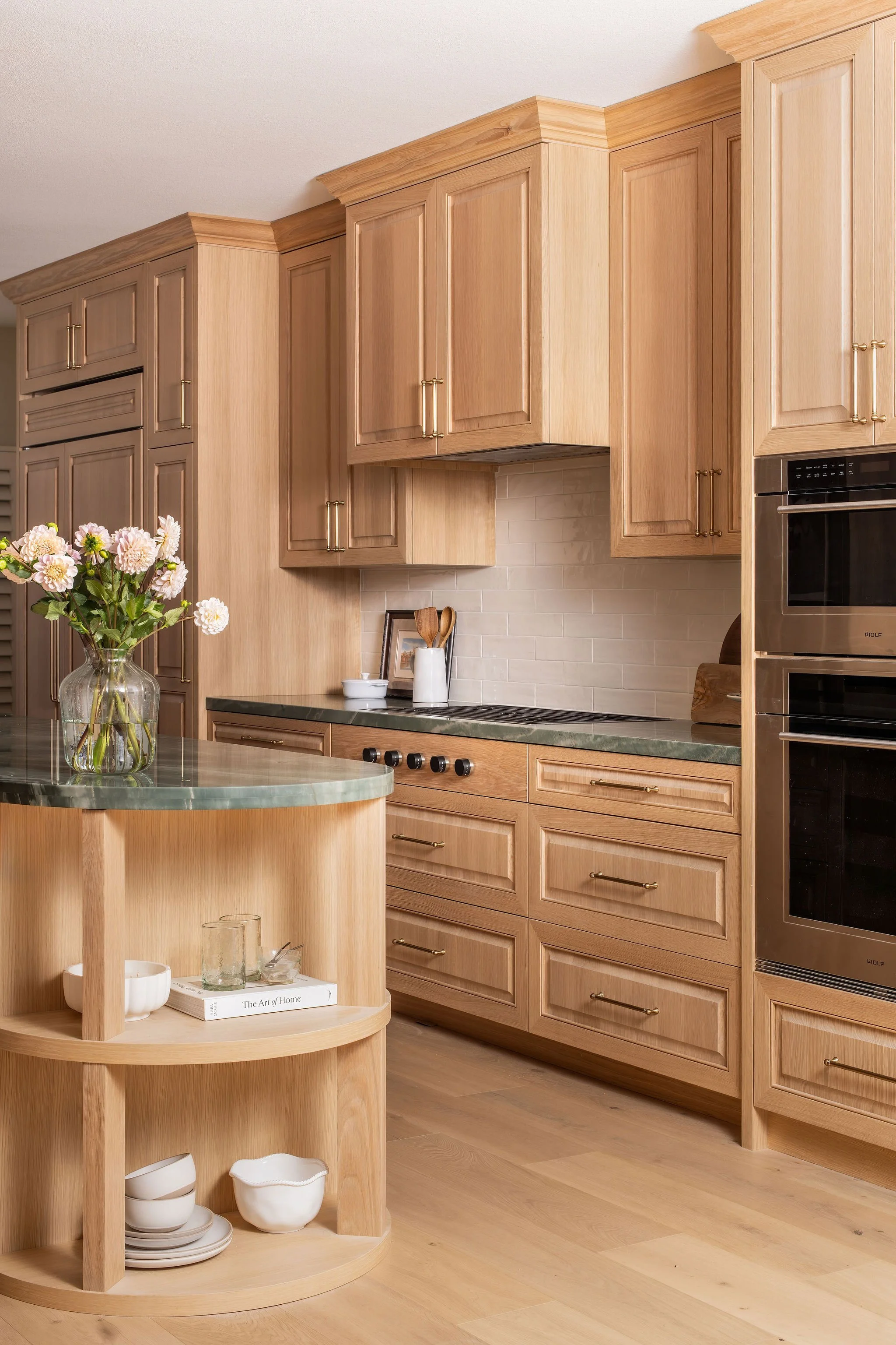 A kitchen with wooden cabinets, a granite countertop, and a round kitchen island with open shelves. A vase with flowers, bowls, and glasses are on the island. There are built-in double ovens on the right side.