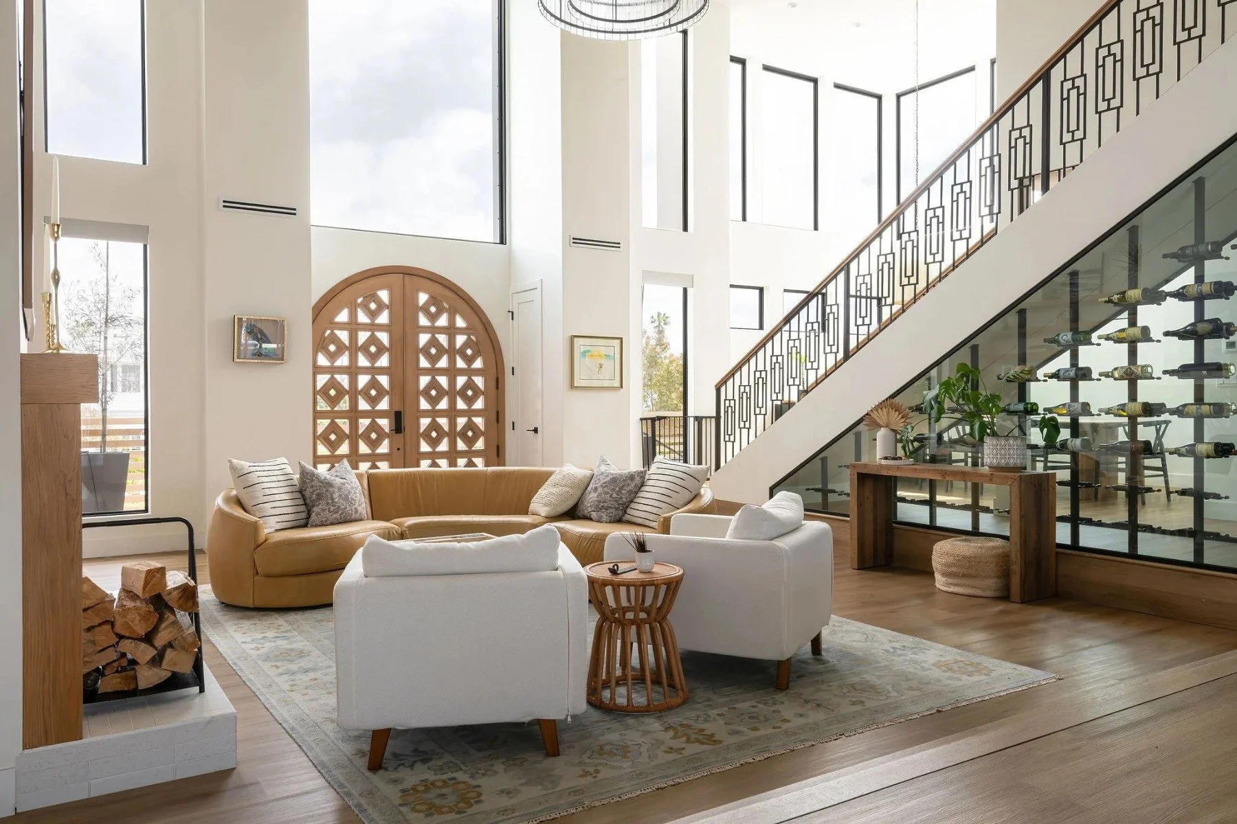 Modern living room with beige and white furniture, wooden accents, large windows, staircase with decorative black railing, and a wine rack on glass wall.