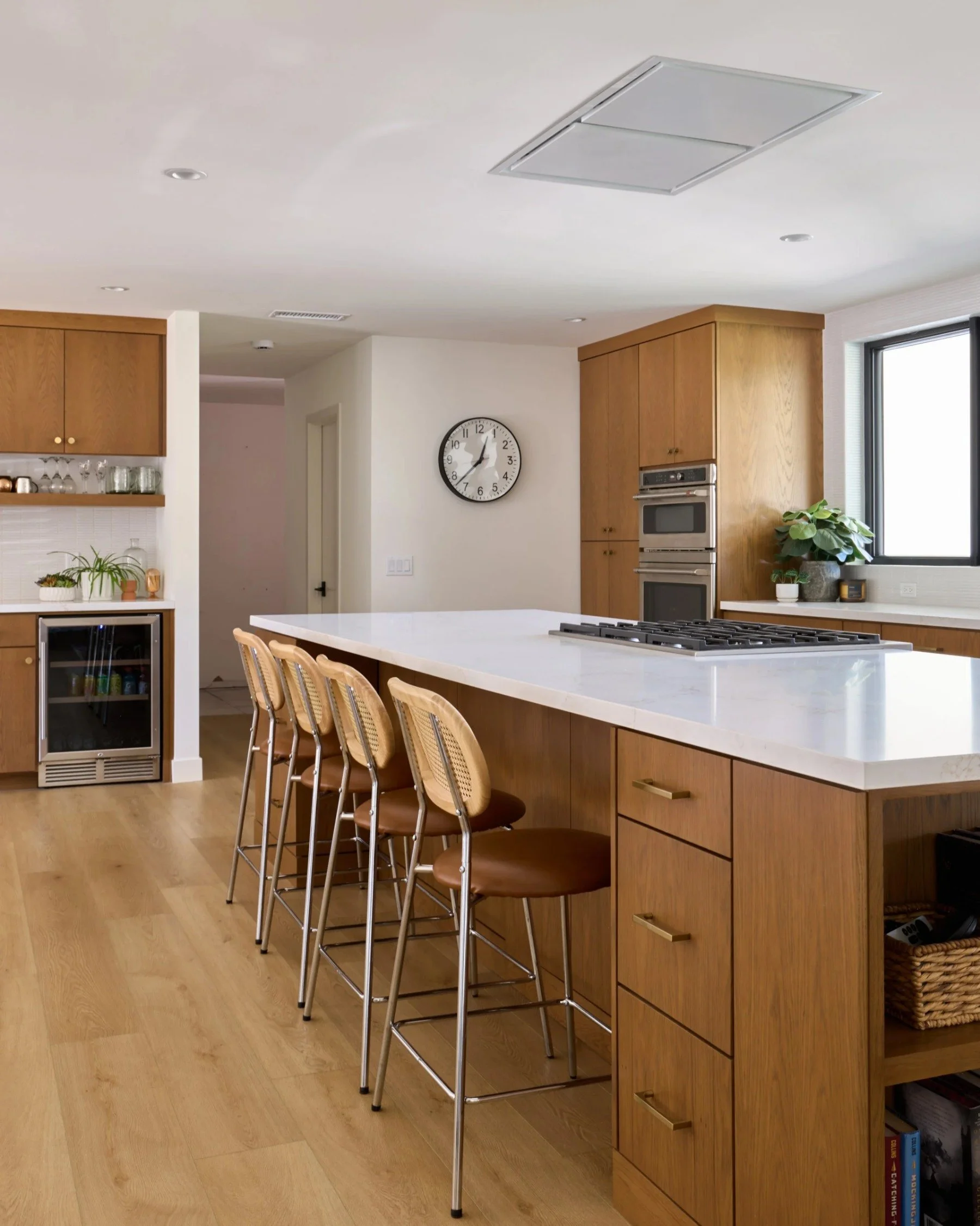 Modern kitchen with a white marble island, four wooden bar stools, oak cabinets, a wall clock, and a window with potted plants.
