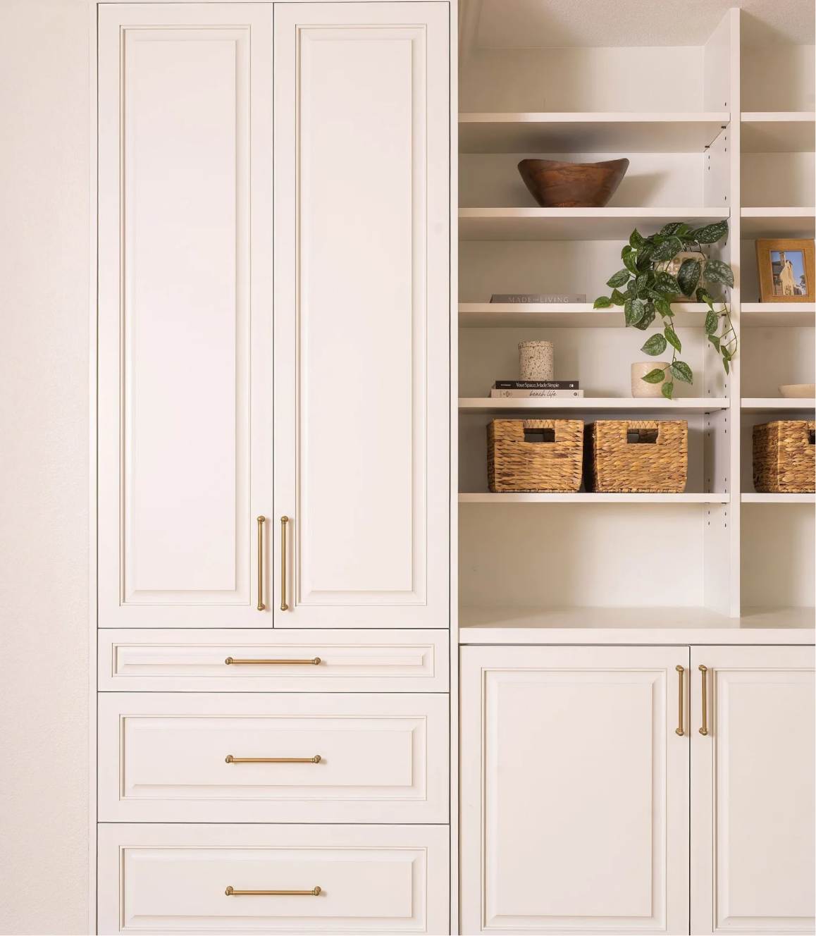 White built-in kitchen or pantry cabinets with gold handles, and open shelves with decorative baskets, a plant, a wooden bowl, and framed photos.