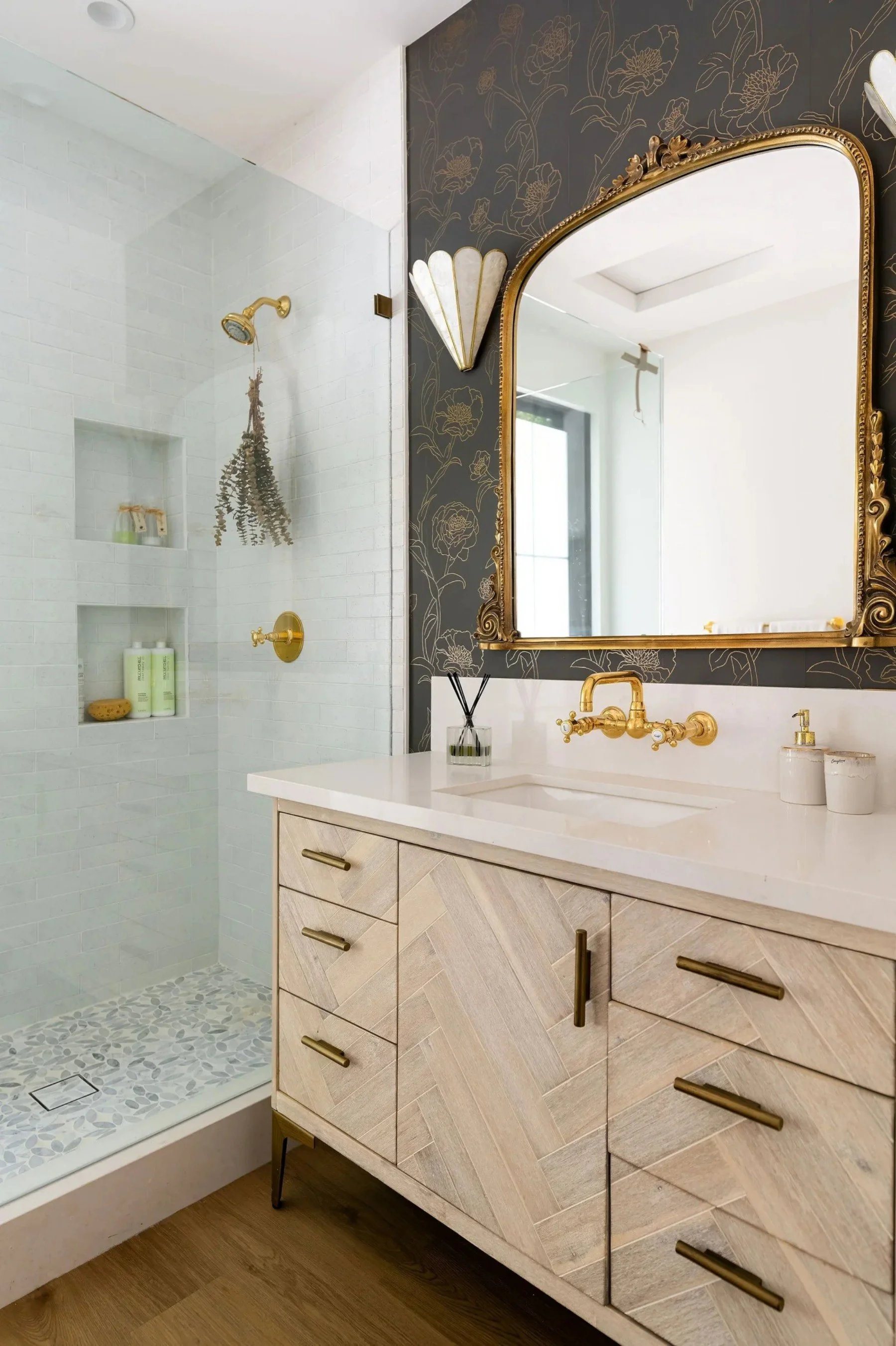 A bathroom with a walk-in shower and a vanity with a decorative mirror. The shower has white tiled walls, built-in shelves with toiletries, and a gold showerhead. The vanity has a light wood cabinet with gold handles, a white countertop, and a gold faucet. The mirror has an ornate gold frame, and a wall sconce with a decorative shade is mounted beside it. The wall behind the mirror features dark wallpaper with a floral pattern.