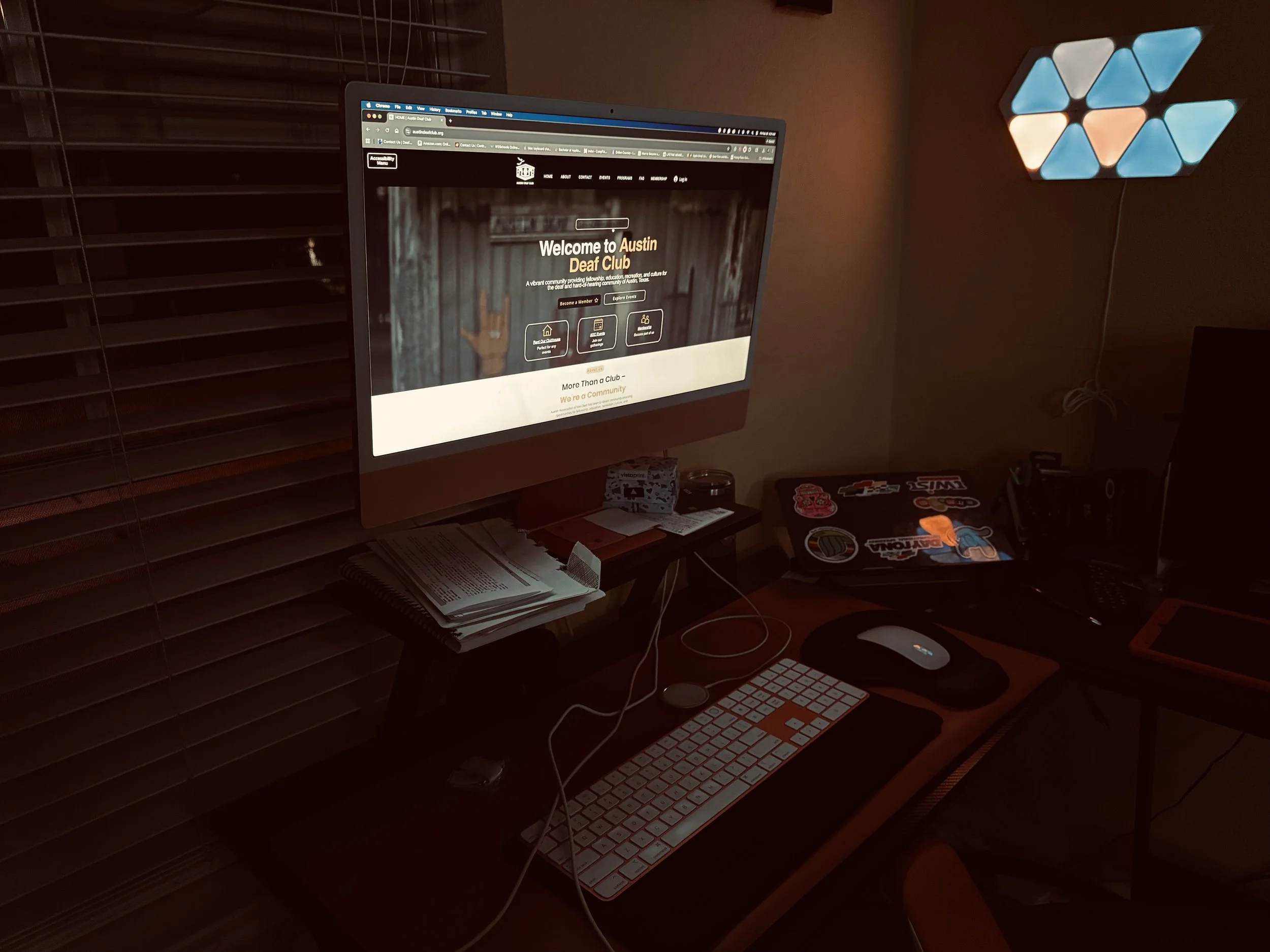 A computer workspace with a monitor displaying the Austin Deaf Club website, a keyboard, mouse, and various stickers on a laptop. There are notebooks and a desk lamp with hexagon-shaped lights on the wall.