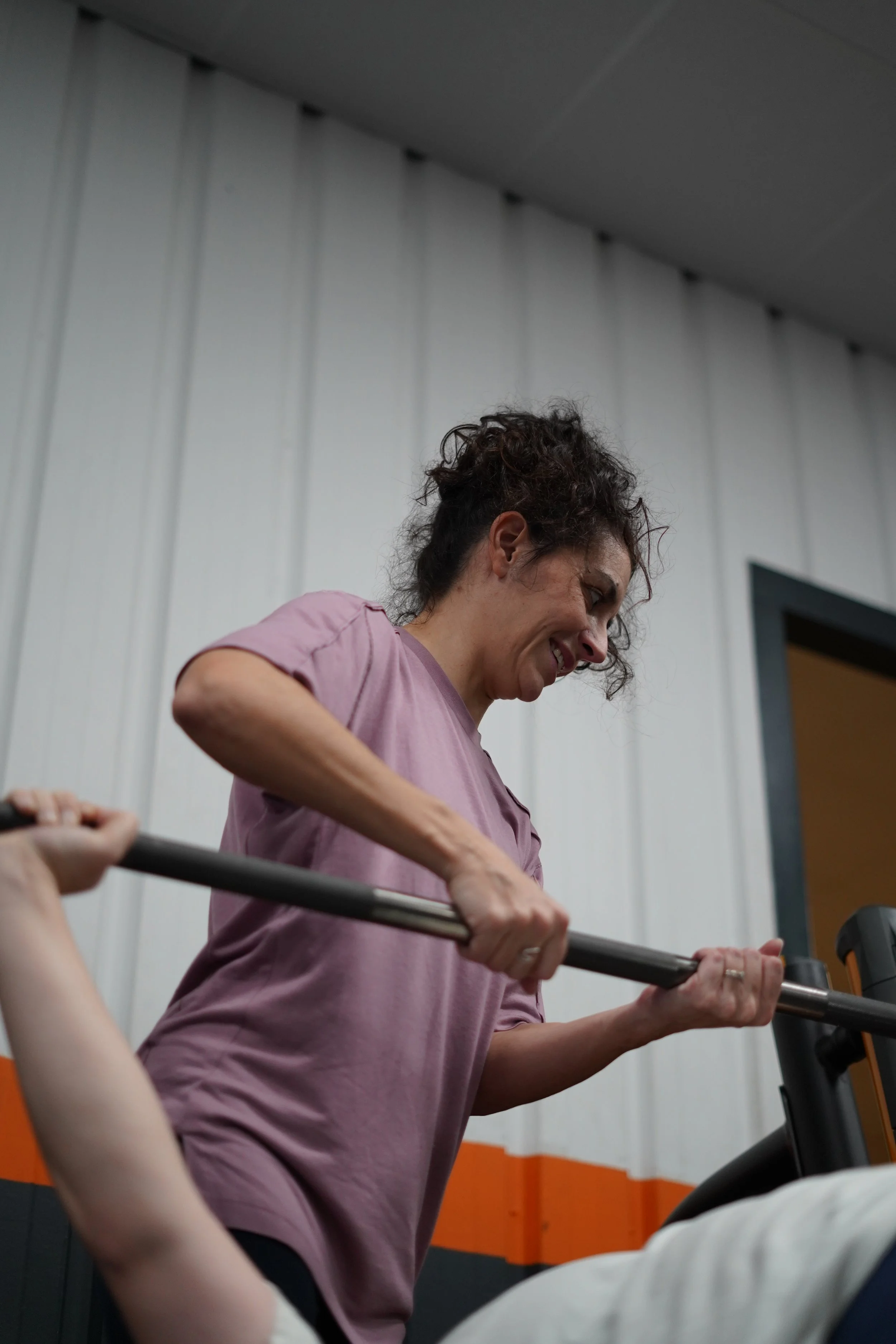 Une femme en tenue de sport effectue une séance de musculation avec une barre dans une salle de sport. Elle porte un t-shirt mauve et a les cheveux bouclés.