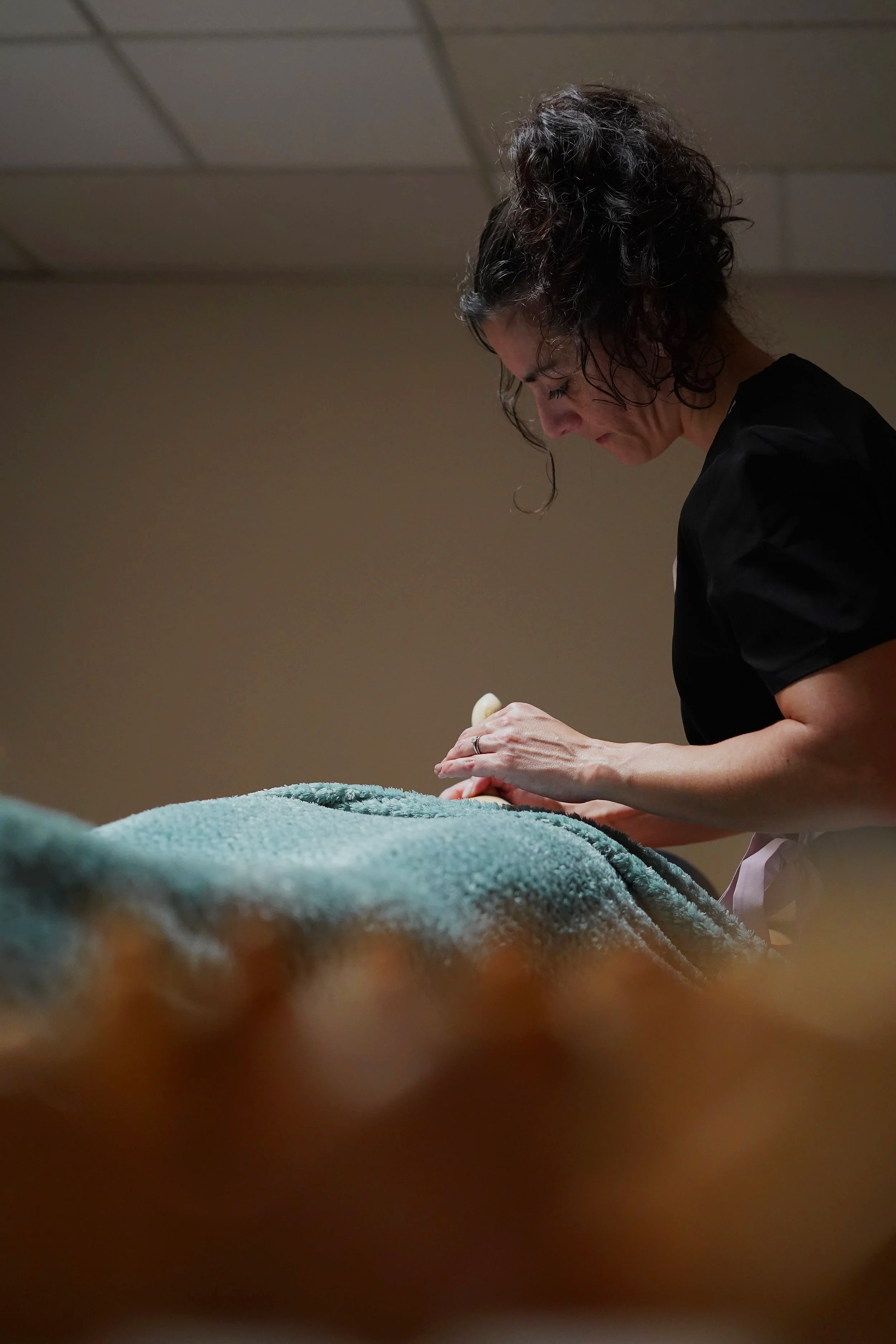 Une femme avec des cheveux bouclés foncés regarde vers le bas, concentrée, probablement lors d'une activité de soin ou de soins personnels, dans une pièce avec plafond de tuiles.