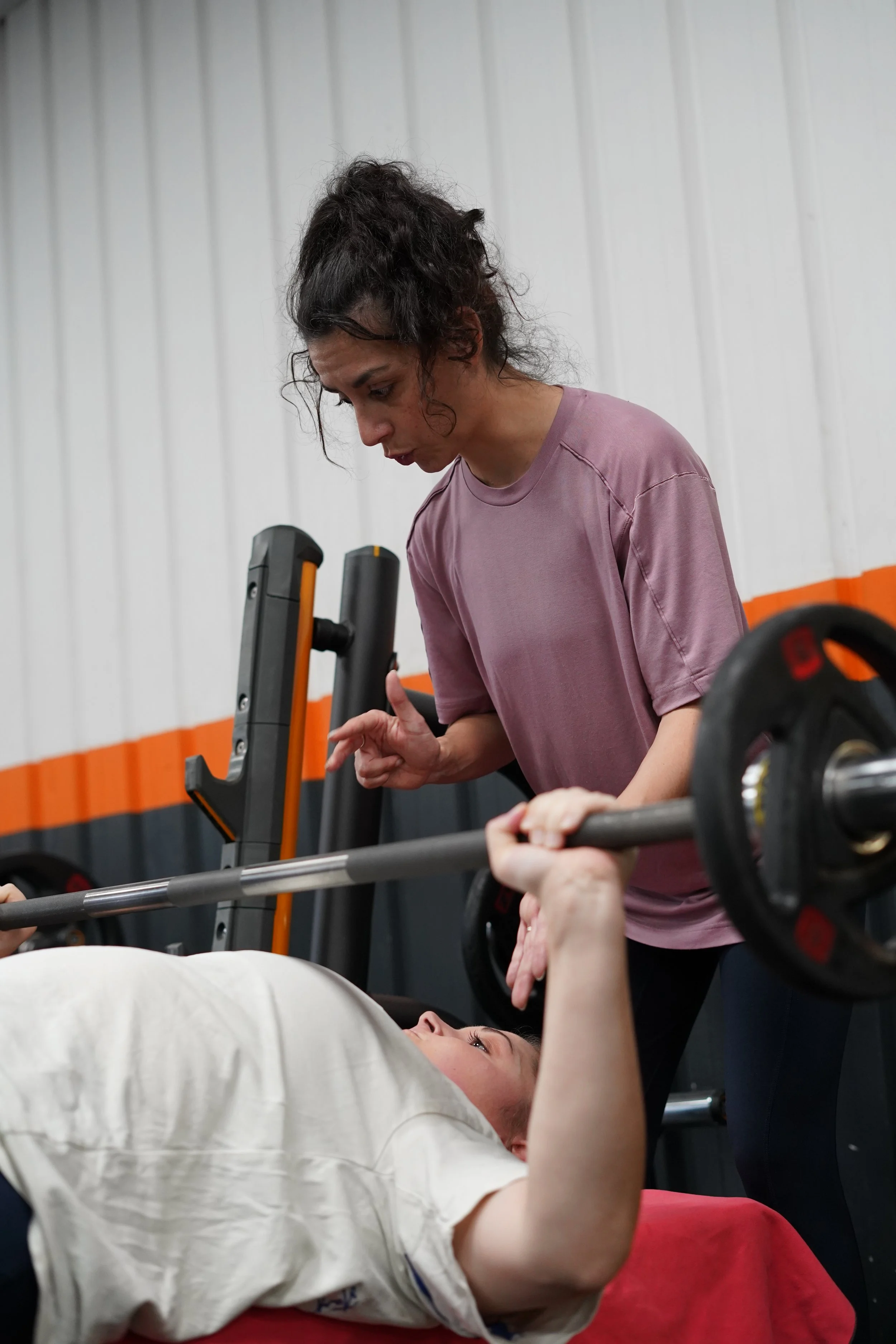 Une femme allongée sur un lit ou une table, tenant un haltère, pendant qu'une autre femme la soutient ou la supervise lors d'une séance d'entraînement en salle de sport.