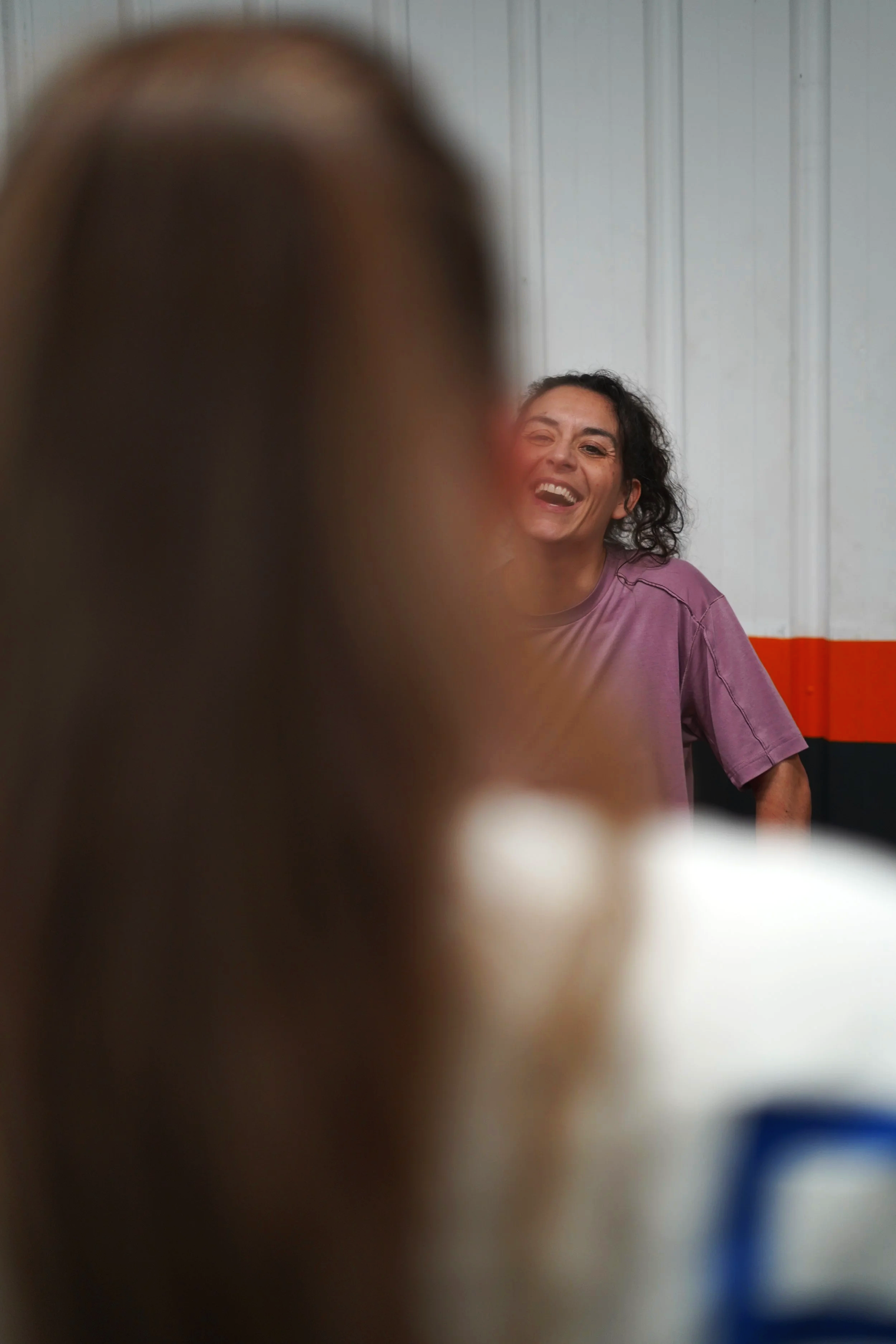 Une femme souriante portant un t-shirt violet, vue en arrière floue, avec un mur blanc en arrière-plan.