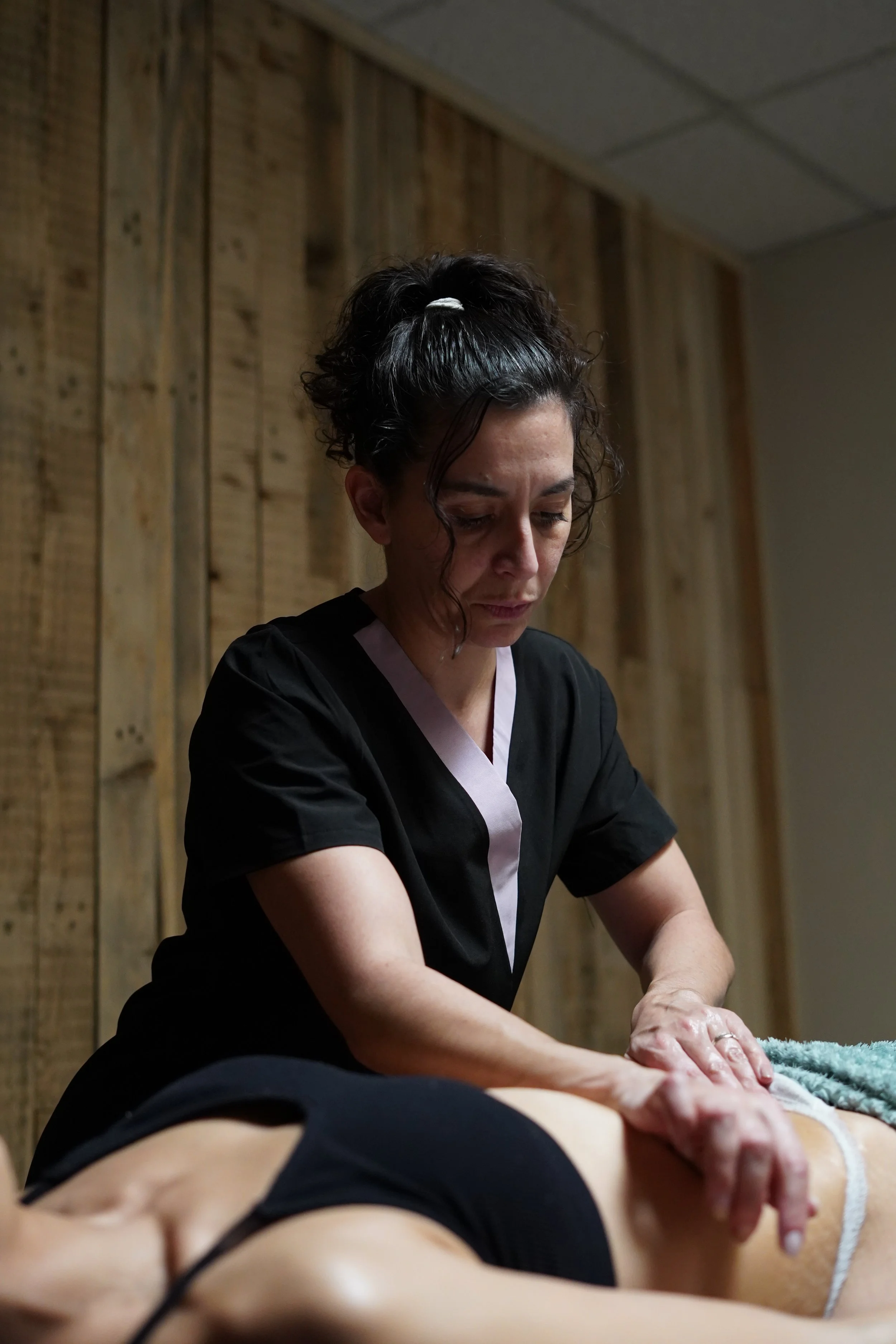 Une femme avec des cheveux foncés coiffés en chignon, portant un uniforme noir avec une bande rose, pratique un massage ou un soin sur une personne allongée. La scène se déroule dans un cadre intérieur avec des murs en bois.