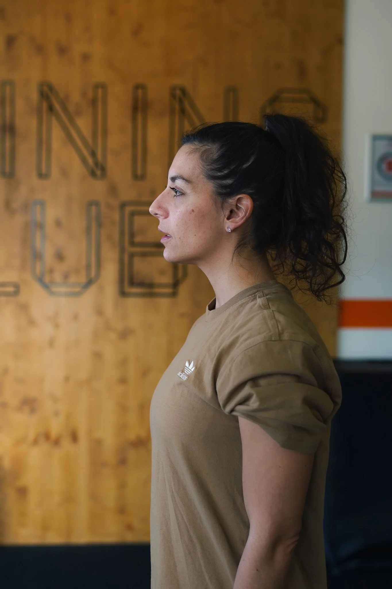Profil d'une femme avec une queue de cheval, portant un t-shirt beige Adidas, se tenant devant un mur en bois avec un logo ou un texte flou.
