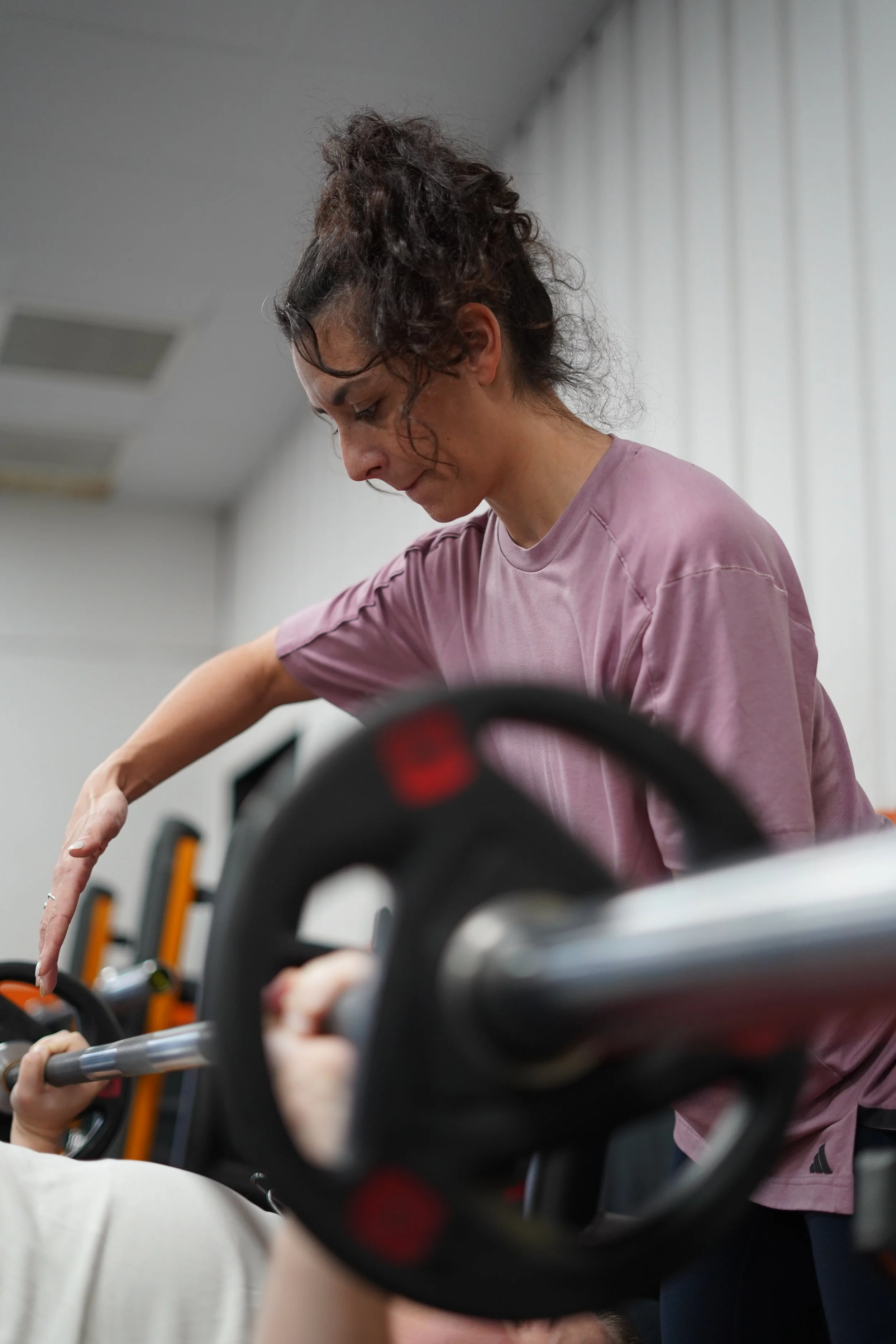 Une femme fait de l'exercice avec une barre de musculation dans une salle de sport, concentration sur son visage, portant un t-shirt rose et des lunettes.