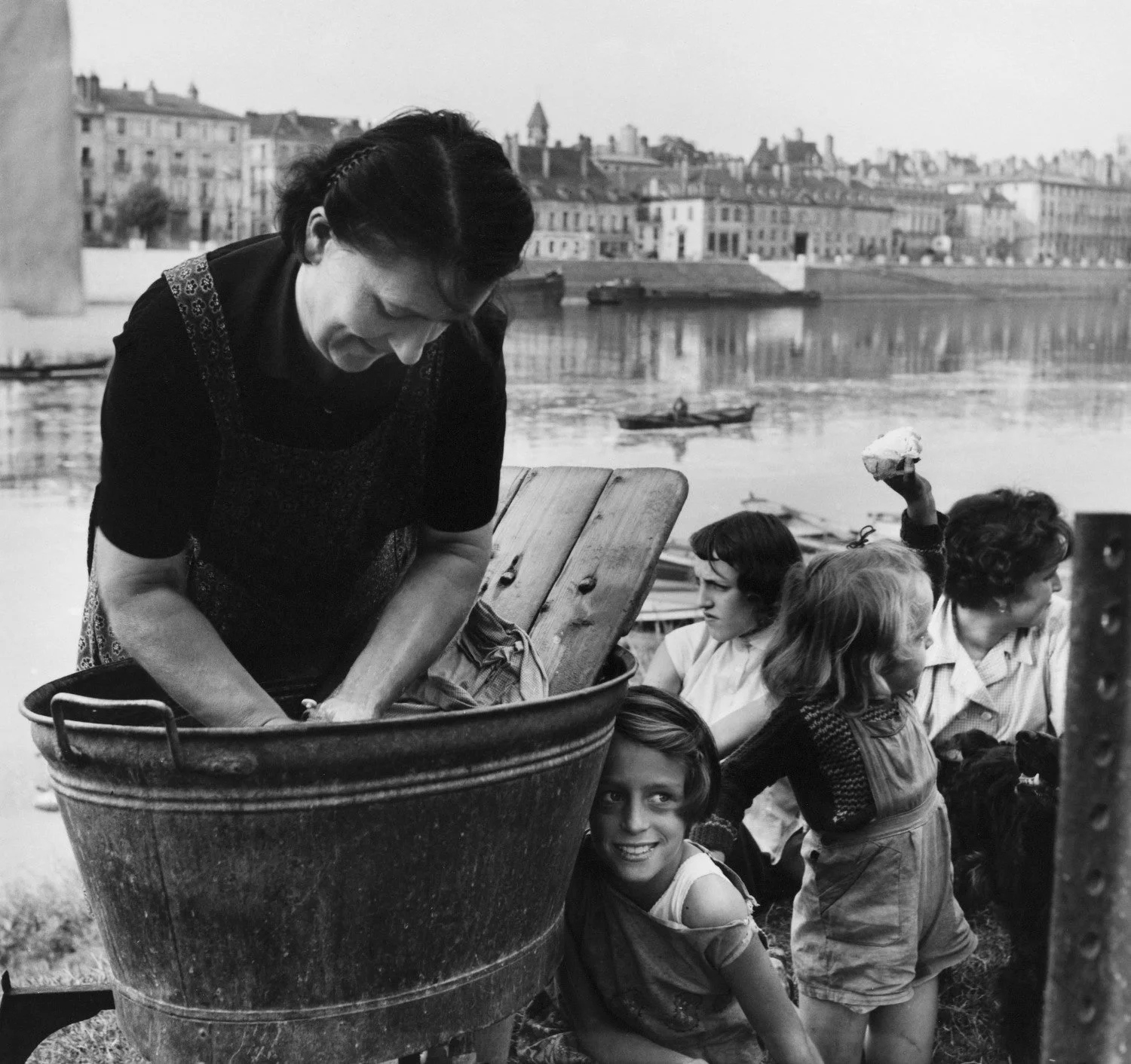 Donna che lava i panni in un tino vicino a un fiume con alcune ragazze sedute e un paesaggio urbano sullo sfondo.
