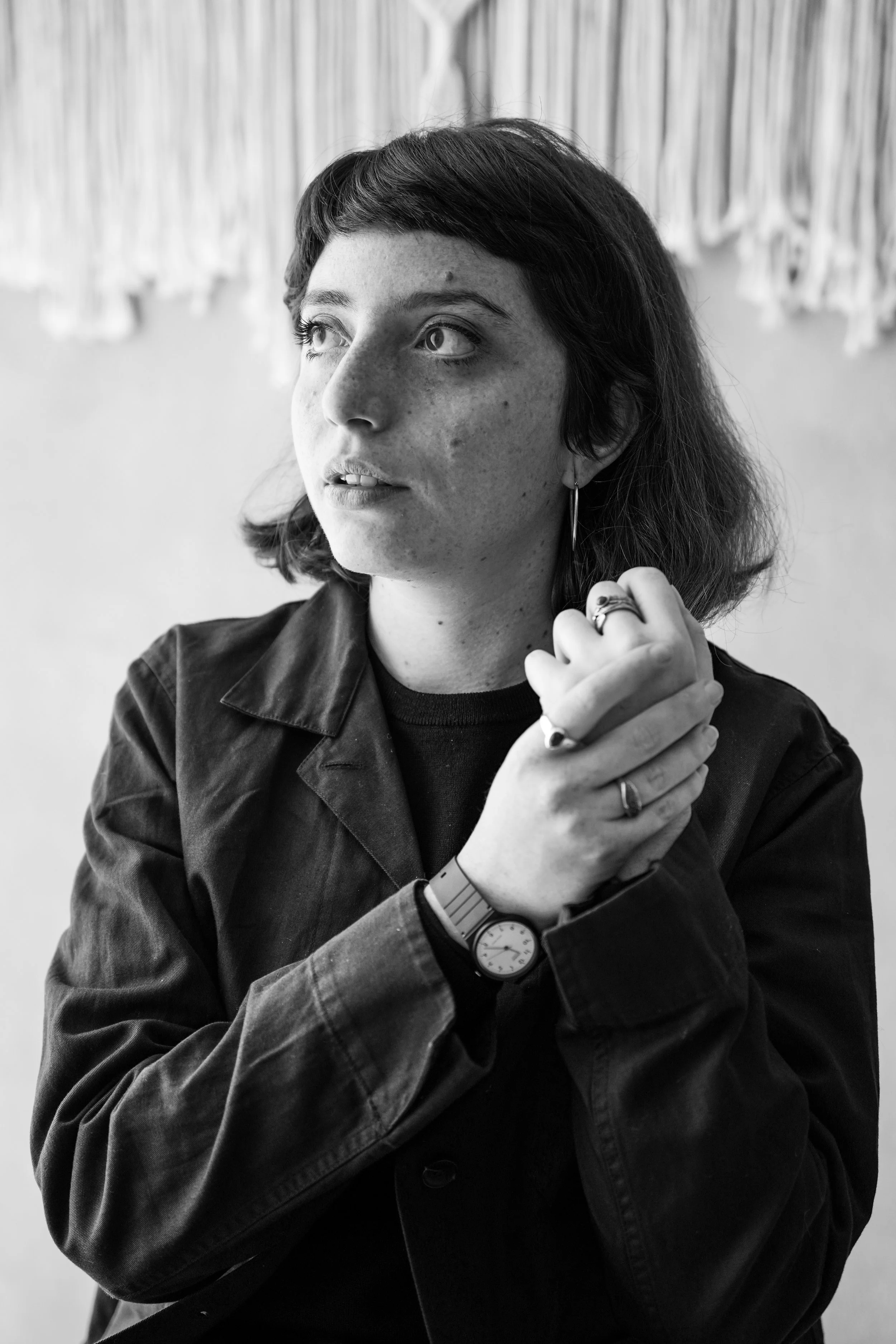 Ritratto in bianco e nero di una donna con capelli corti, giacca di pelle, oppure di jeans, che guarda verso sinistra con espressione pensierosa, con anelli, orecchini, orologio e braccialetto, sullo sfondo una parete decorata.