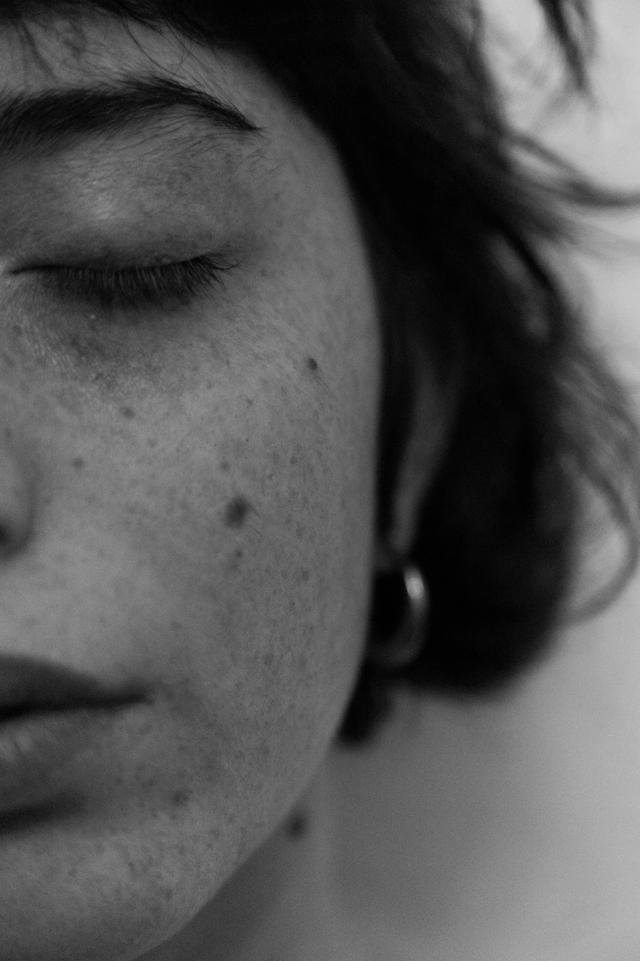 Primo piano di una persona con occhi chiusi, pelle con macchie e lentiggini, e orecchino, in nero e bianco.