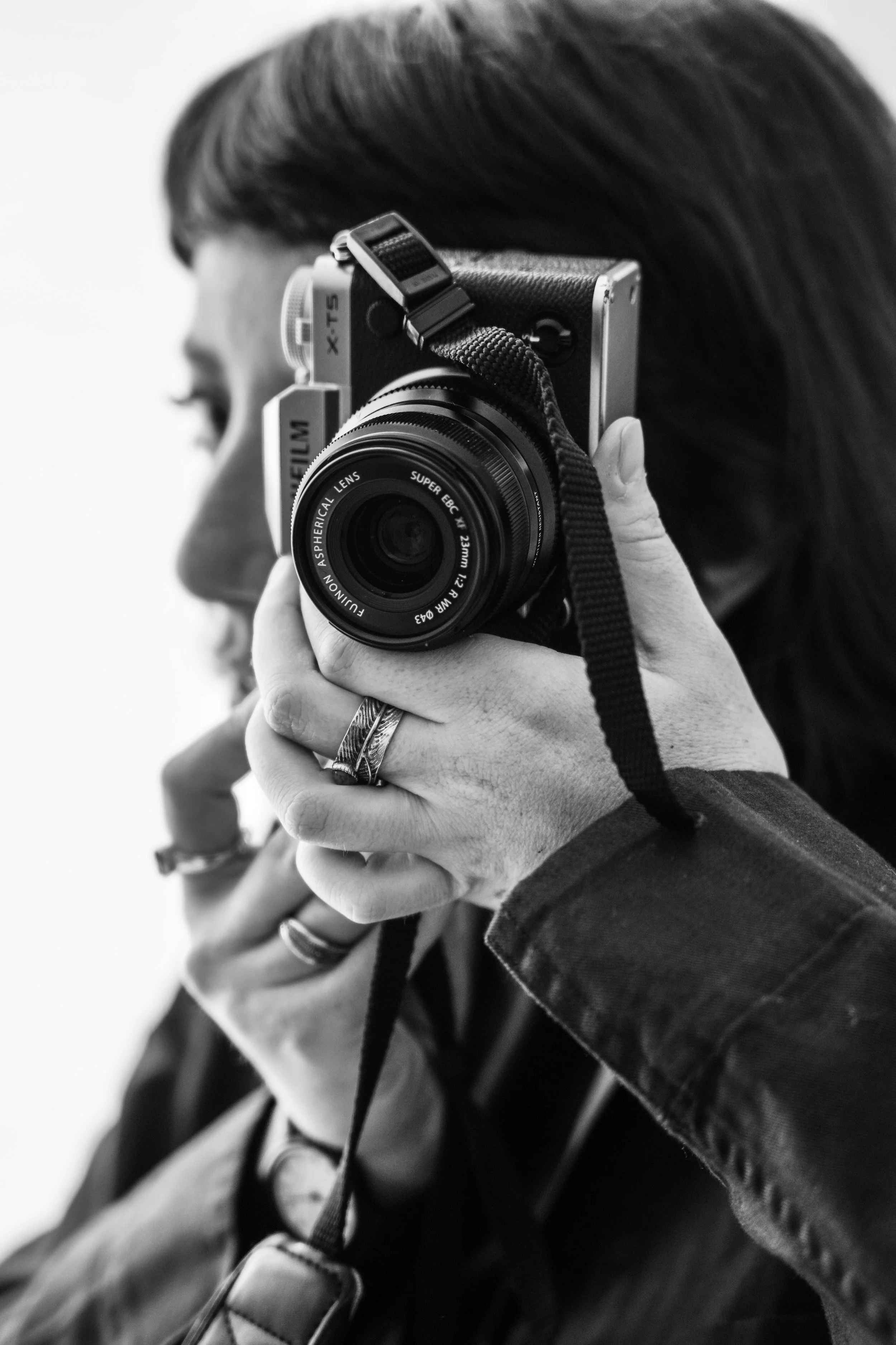 Close-up di una persona con capelli scuri che tiene una fotocamera reflex digitale davanti a sé, visibile il dettaglio del obiettivo e delle mani.