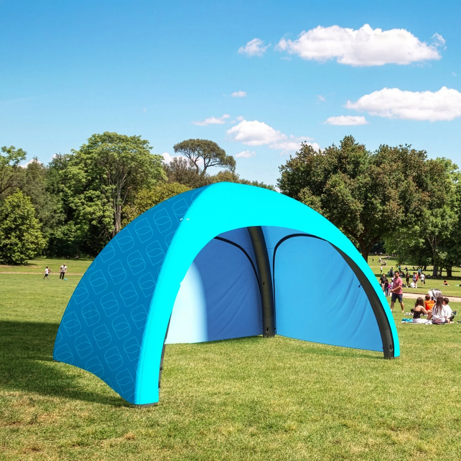 Tenda pubblicitaria blu in un parco con persone che si rilassano e giocano tra gli alberi e il prato verde.