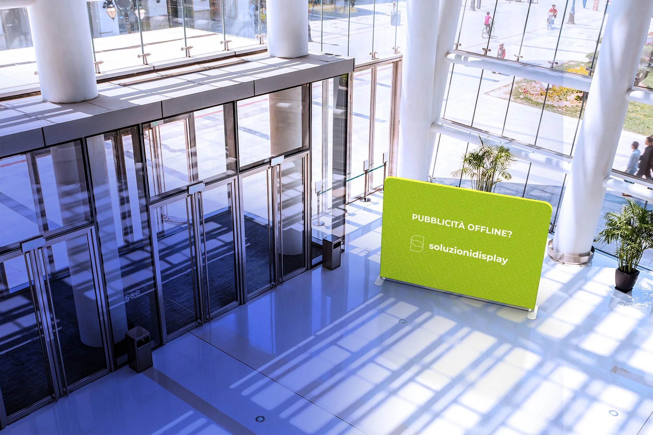 Interno di un edificio moderno molto lumino con grandi vetrate e piante, con una parete pubbicitaria verde con la scritta scritto "Pubblicità offline? soluzionidisplay".